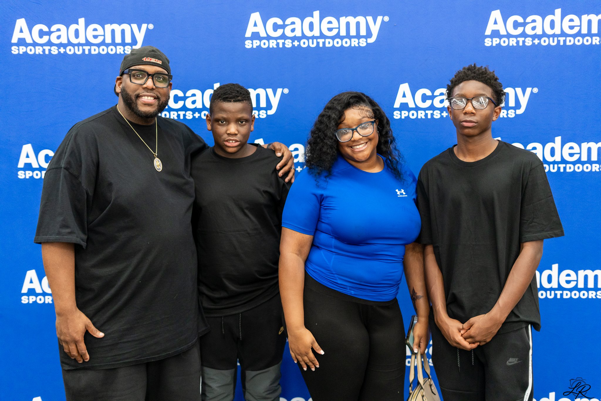 Four people standing in front of a blue backdrop with white text that reads "Academy Sports+Outdoors". The group includes two men and two women, all wearing casual sportswear and glasses.