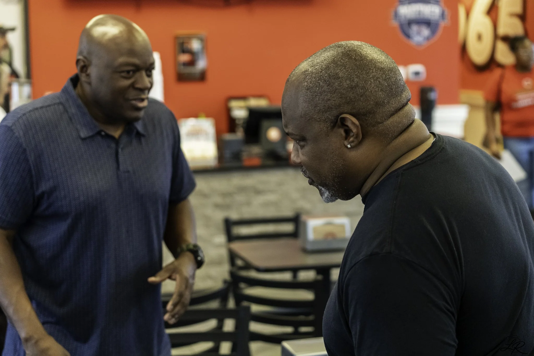 Two men are engaged in a conversation inside a red-walled restaurant or cafe, with one man standing and the other sitting. The standing man is wearing a blue short-sleeved shirt, and the sitting man has a close-cropped hairstyle and is looking down. 