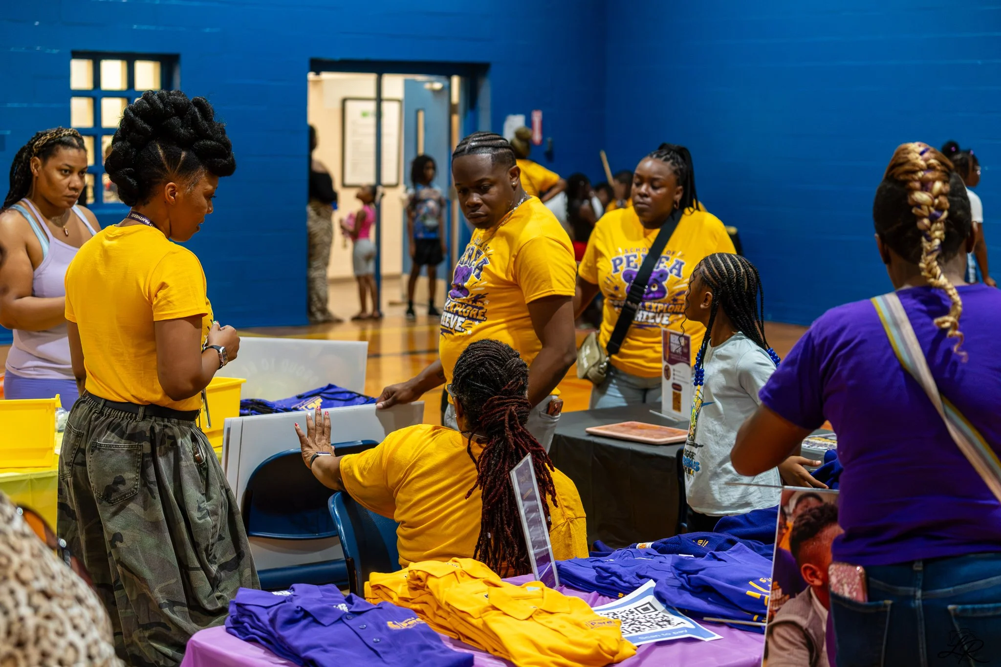 People gathered at a table with yellow and purple t-shirts, engaging in a community event inside a gymnasium with blue walls.