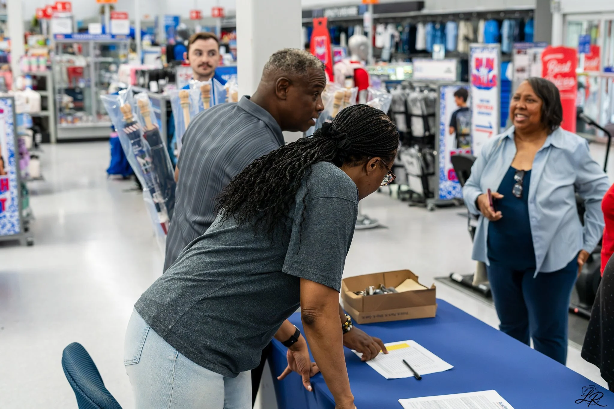 A man with short gray curly hair and a woman with long braided hair leaning over a table, signing documents in a retail store. An employee is smiling and talking to them in the background, while shelves of various products are visible in the store.