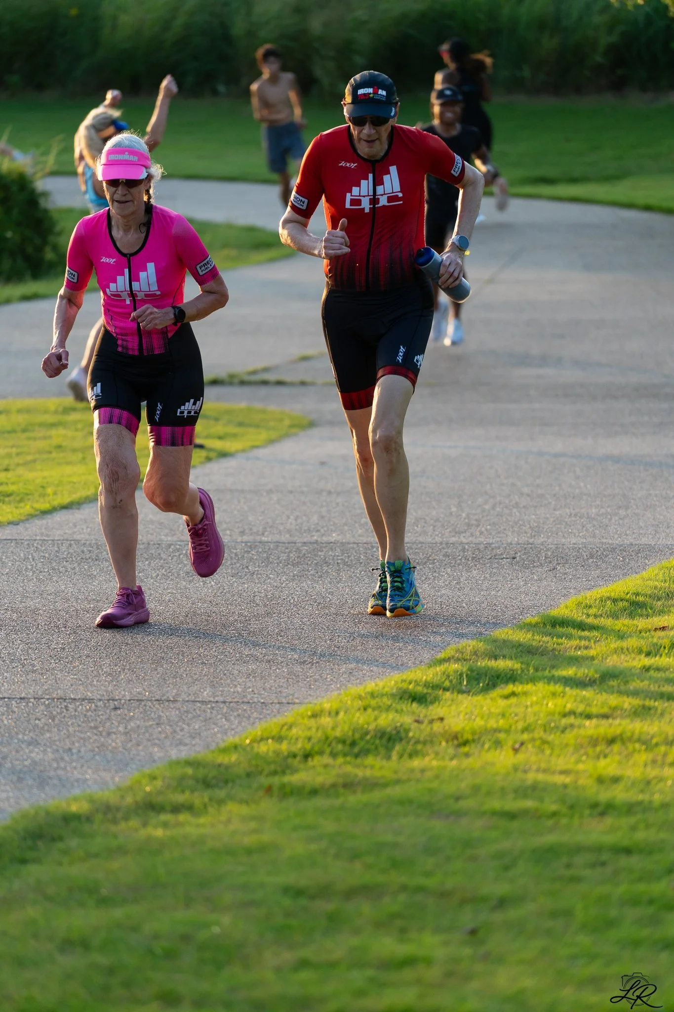 Older man and woman running on a paved trail in a park, surrounded by grass and trees, during sunset.
