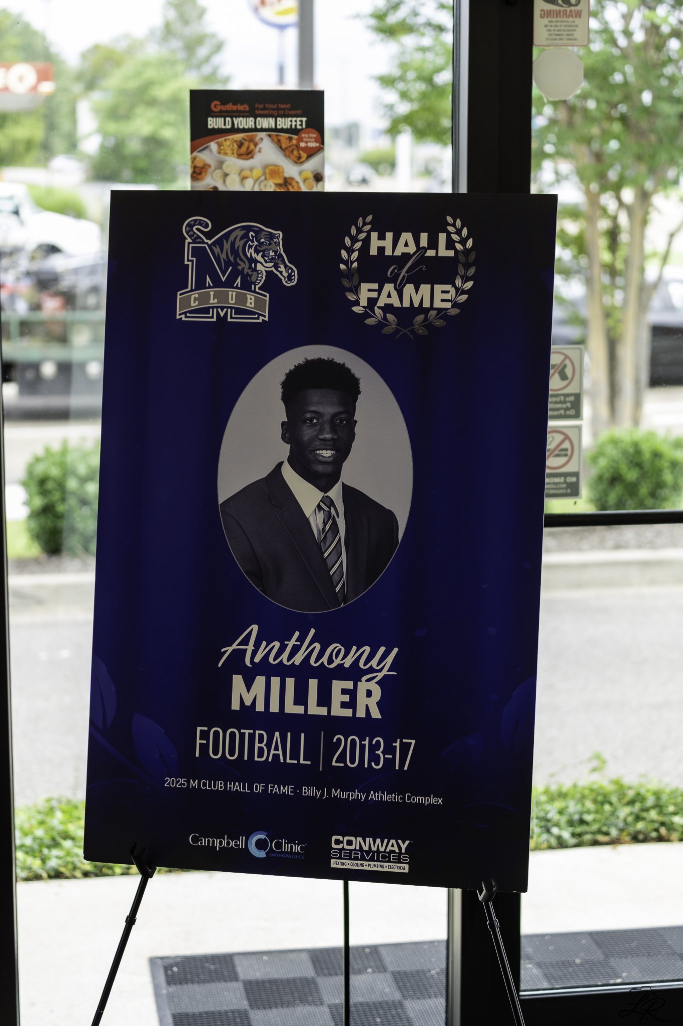 A blue poster board displayed on a stand honors Anthony Miller as a member of the football hall of fame 2013-17. The poster features a black and white portrait of Anthony Miller in a suit, the hall of fame logo, and logos for Campbell Clinic and Conw