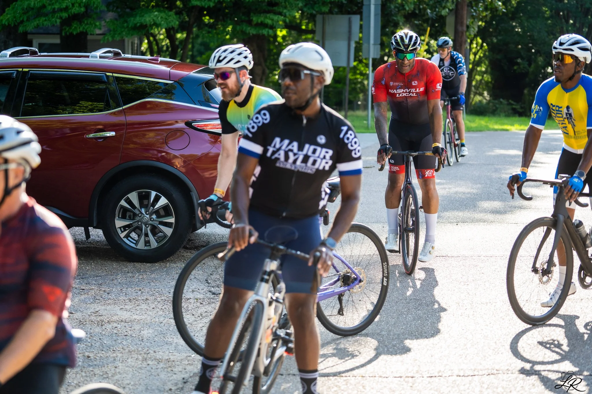 Cyclists in colorful jerseys and helmets riding bikes on a tree-lined street.