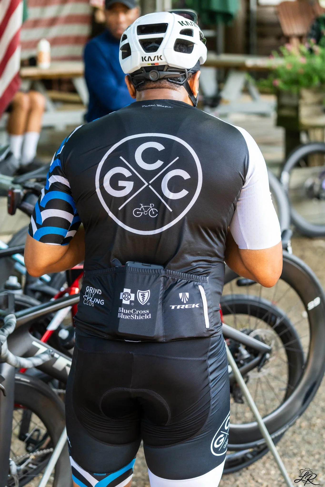 Person wearing a black cycling jersey with logos, a white helmet, and black cycling shorts with logos, standing next to bicycles. The focus is on the back of the cyclist, with a person sitting at a table blurred in the background.