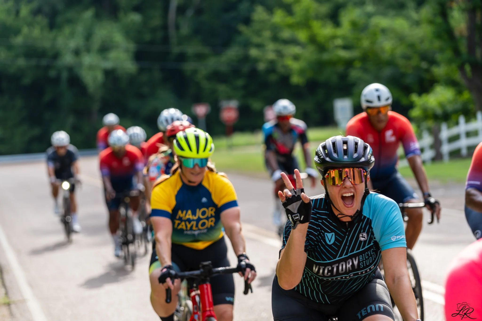 A group of cyclists, wearing safety helmets and colorful sport jerseys, riding on a road surrounded by green trees. They appear to be participating in a cycling event or race, with some smiling and making peace signs.