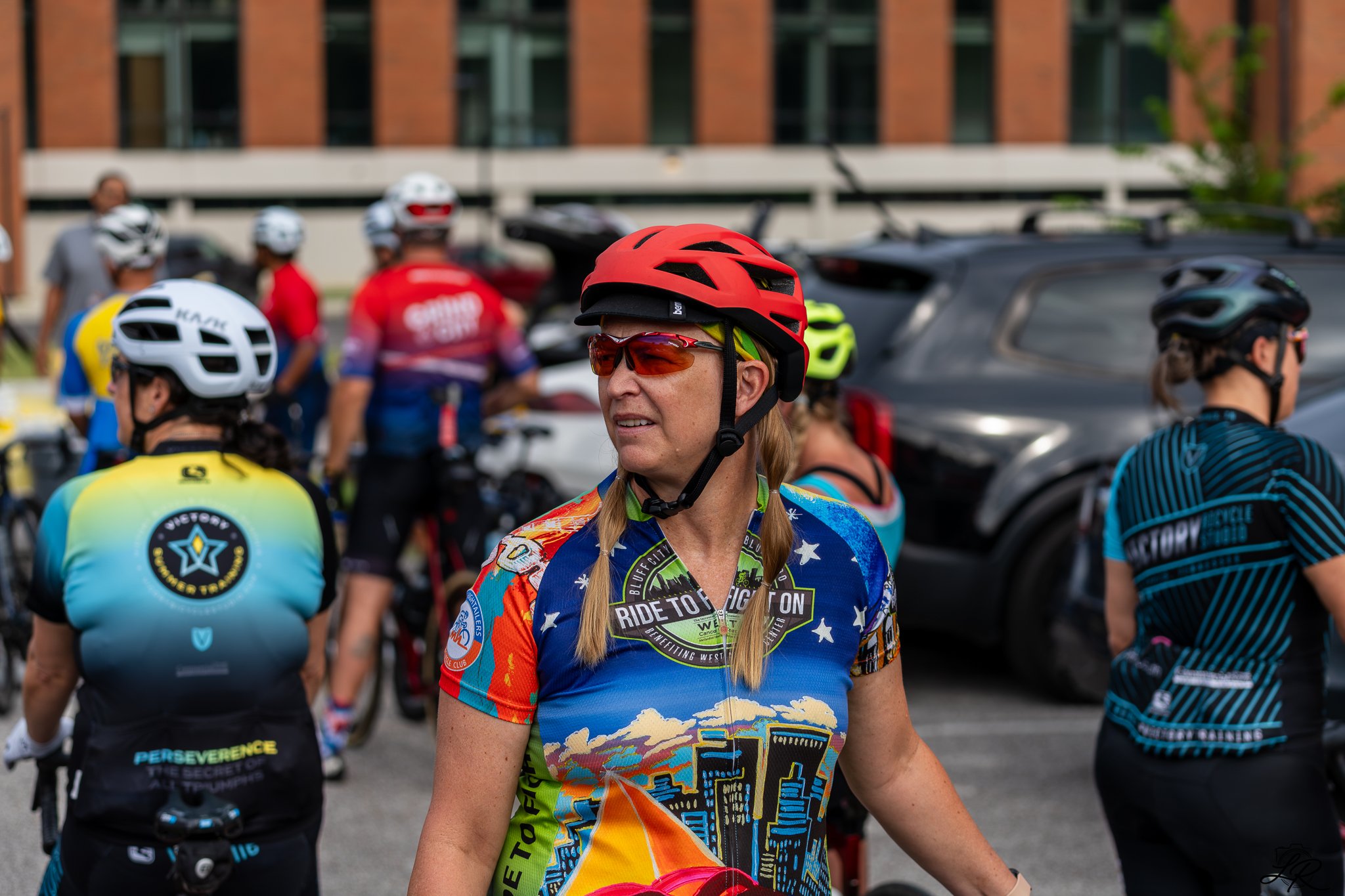 A group of cyclists wearing colorful jerseys and helmets gathers outdoors in front of parked cars, with a woman with long braids, wearing a red helmet and sunglasses, standing in the foreground.