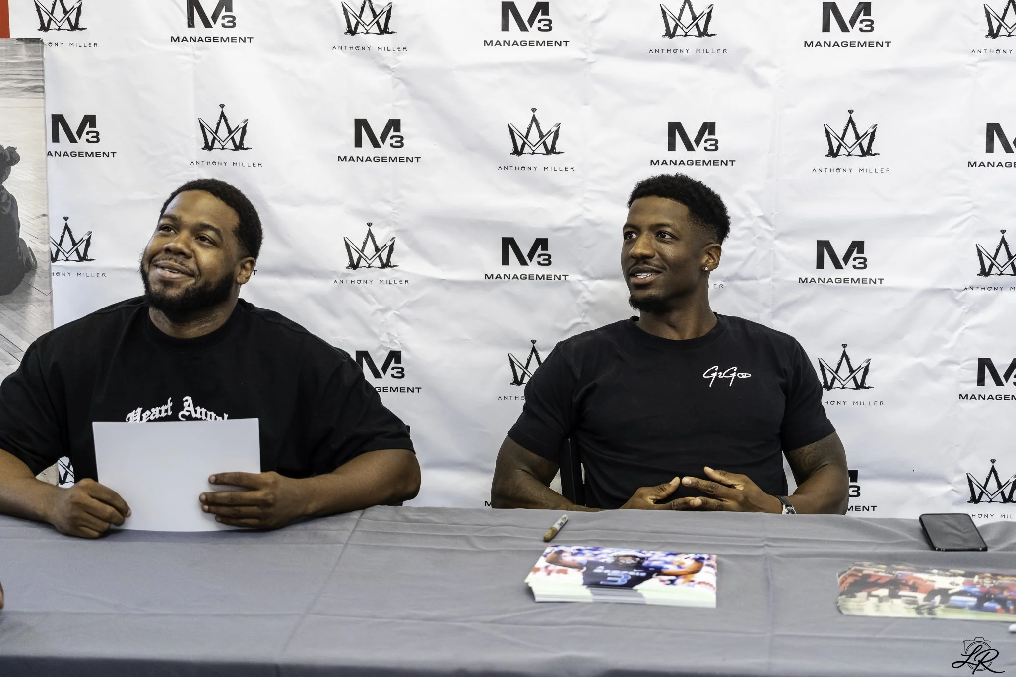 Two men sitting at a table during an event with a white backdrop featuring logos for 'M3 Management' and 'Anthony Miller'. One man is smiling and holding a piece of paper, the other man is looking to his right.
