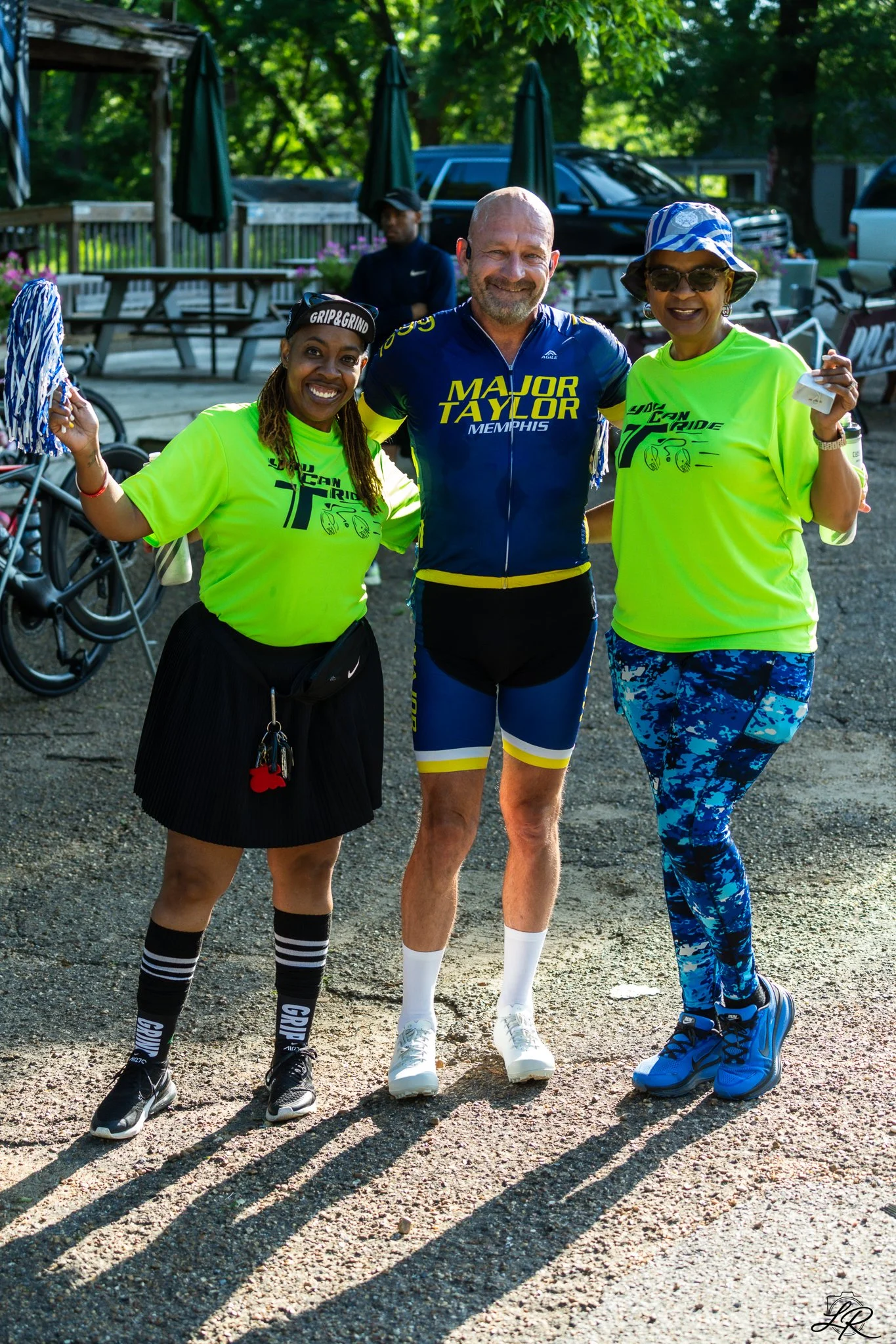 Three people disguised as runners smiling outdoors with trees, parked cars, picnic tables, and umbrellas in the background.