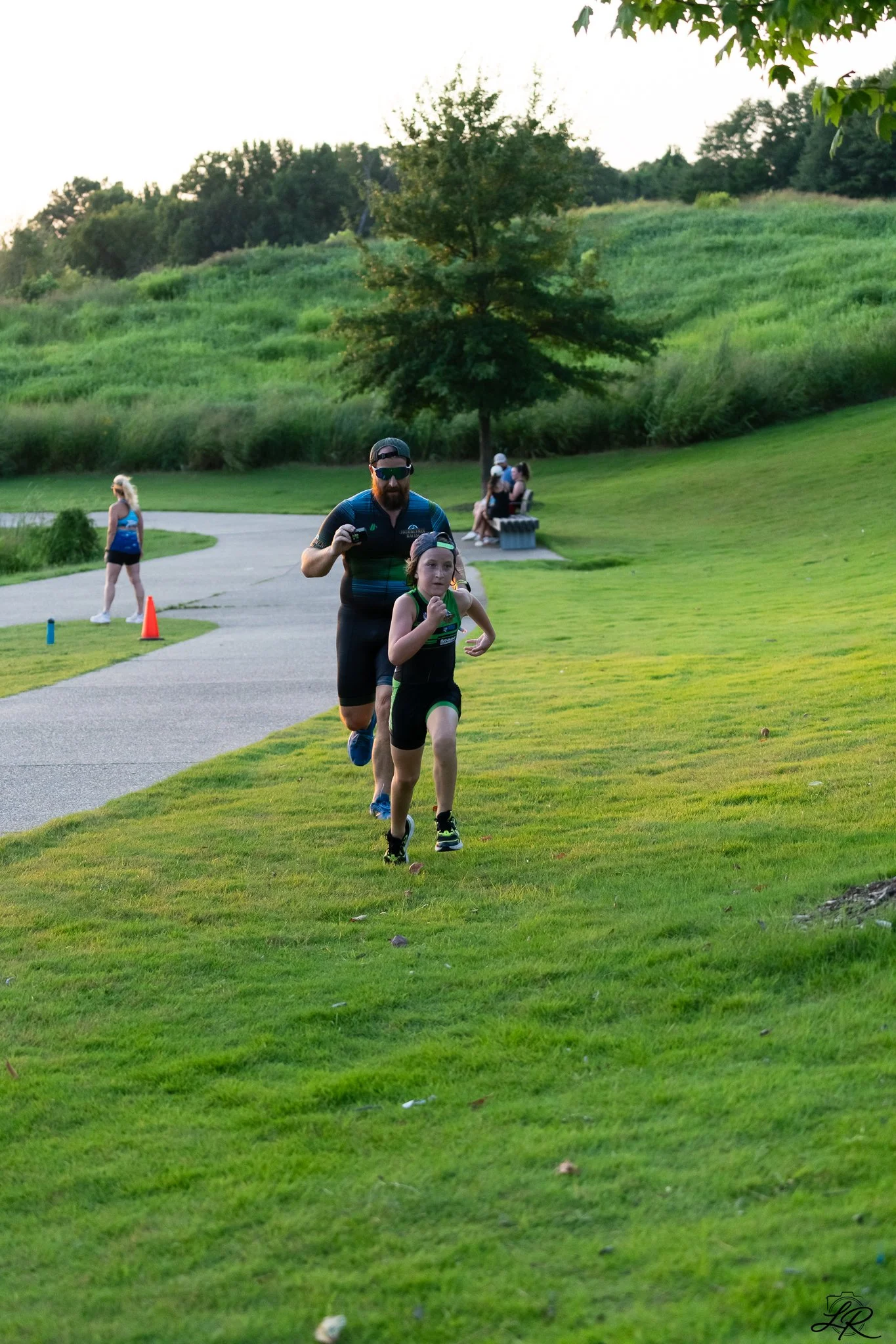 A man and young girl are running on a grassy field in a park. The man is wearing a blue athletic shirt, black shorts, sunglasses, and a hat. The girl is wearing a black sleeveless top and shorts, and a cap. In the background, there are benches with p