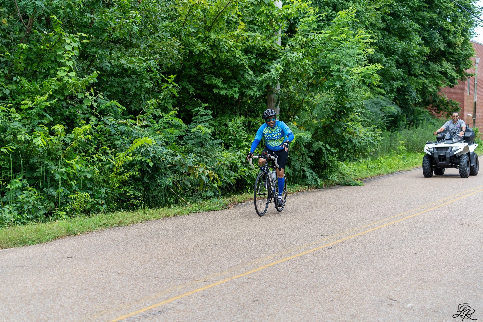 A cyclist in a blue and yellow cycling outfit with a helmet riding on the side of a road next to dense green foliage. An off-road vehicle is following behind on the road, and a brick building is visible in the background.