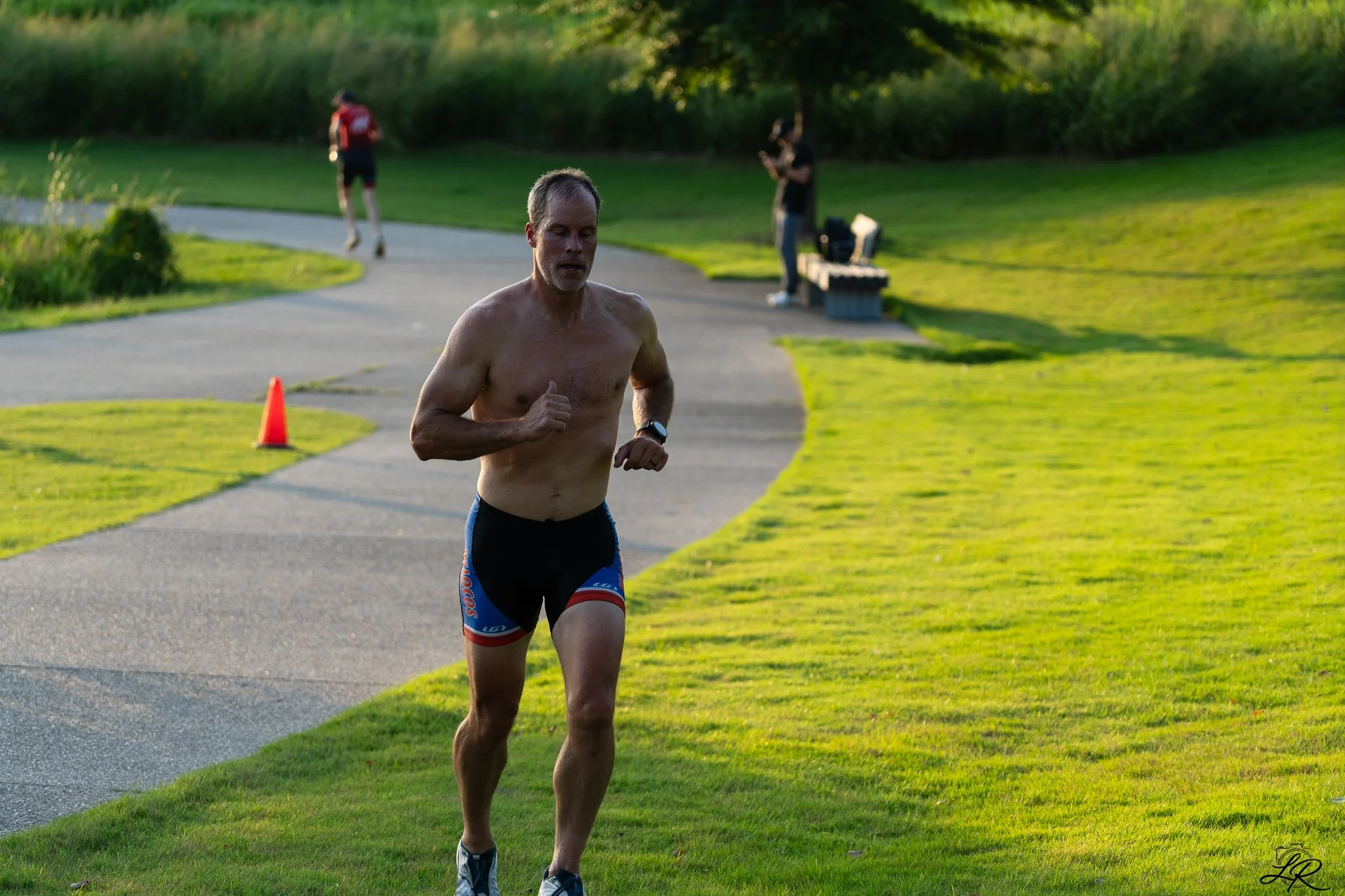 A shirtless man runs on a pavement path in a park, with two other people in the background, one on a bench and another on the path, surrounded by green grass and trees.