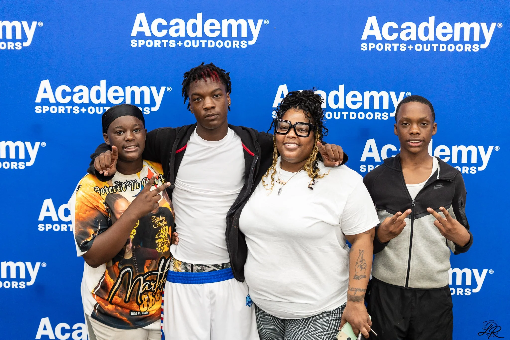 A group of five people standing in front of a blue backdrop with the text 'Academy Sports + Outdoors' printed on it. They are smiling, posing for the photo, and making peace signs with their hands.