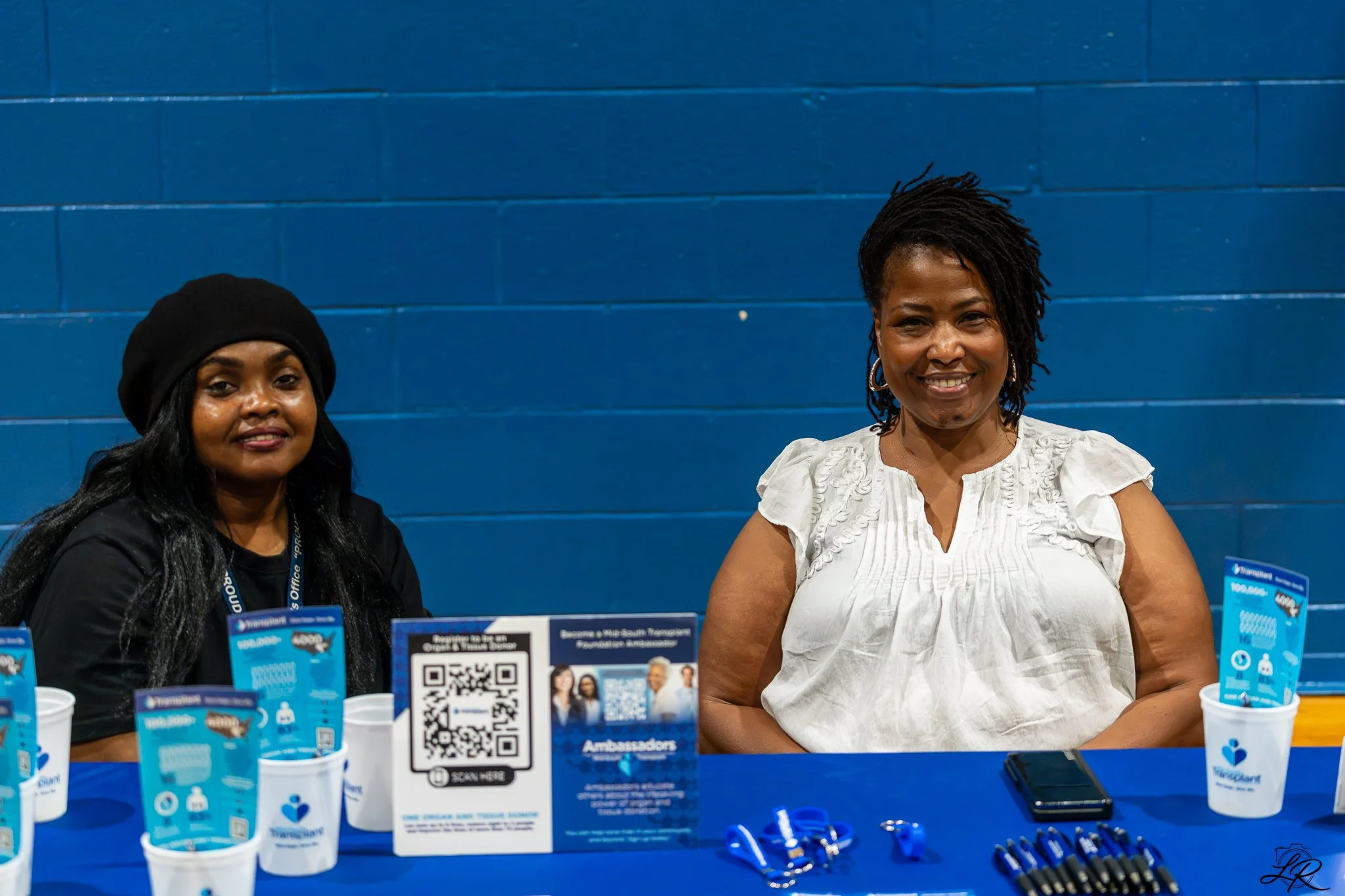 Two women are smiling and sitting at a table with promotional materials in front of a blue wall. The woman on the left wears a black hat and dark clothing, while the woman on the right has short black hair styled in dreads and is dressed in a white t