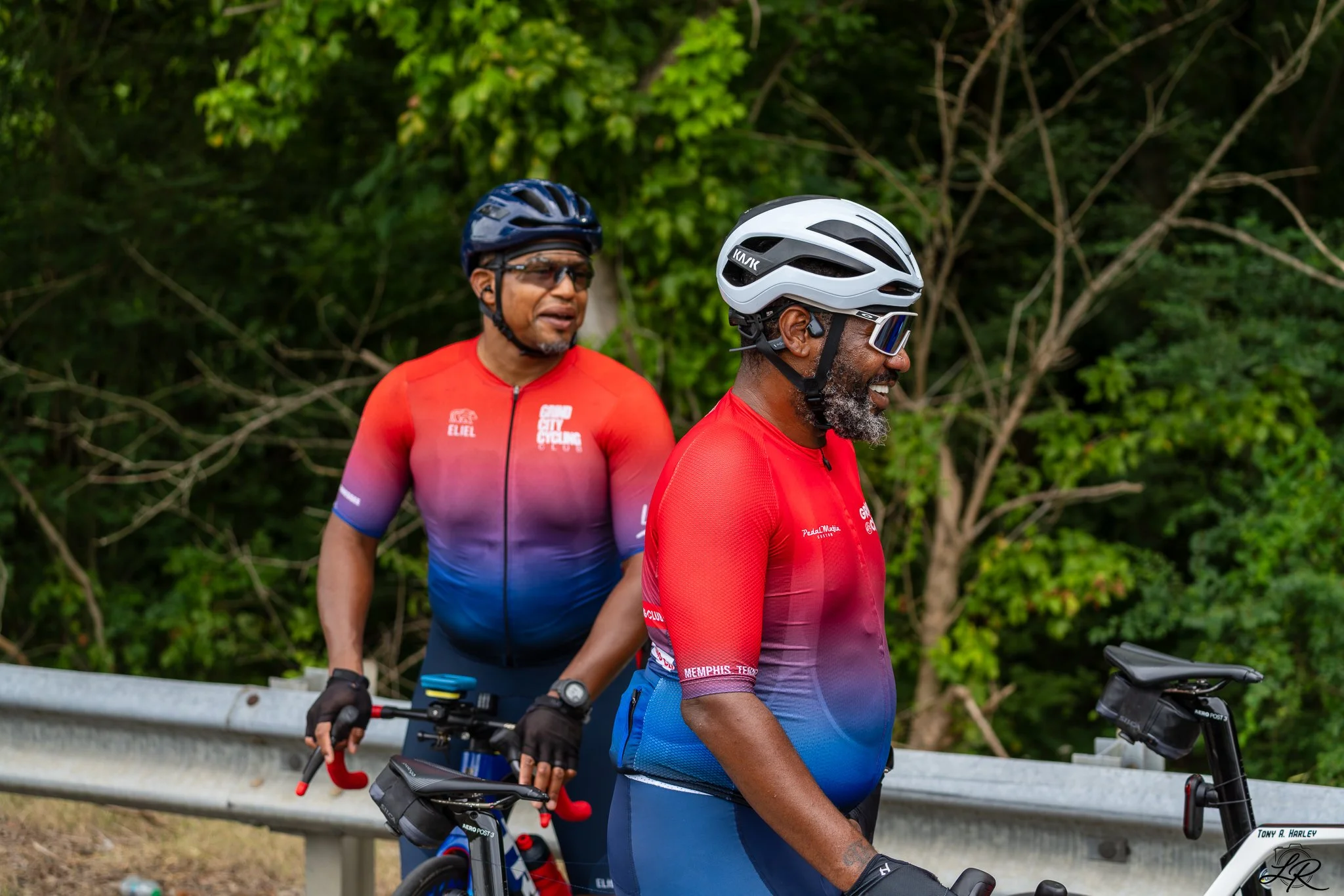 Two men riding mountain bikes on a trail with green trees and bushes in the background. Both men are wearing helmets and cycling gear, and the man in the foreground has a gray beard and reflective sunglasses.