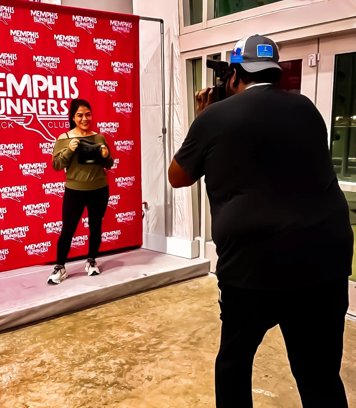 A woman standing in front of a red backdrop with Memphis Runners branding, holding a black purse, smiling, as a photographer takes her picture. The photographer is wearing a black shirt and a baseball cap. The scene appears to be at an indoor event or photo shoot.