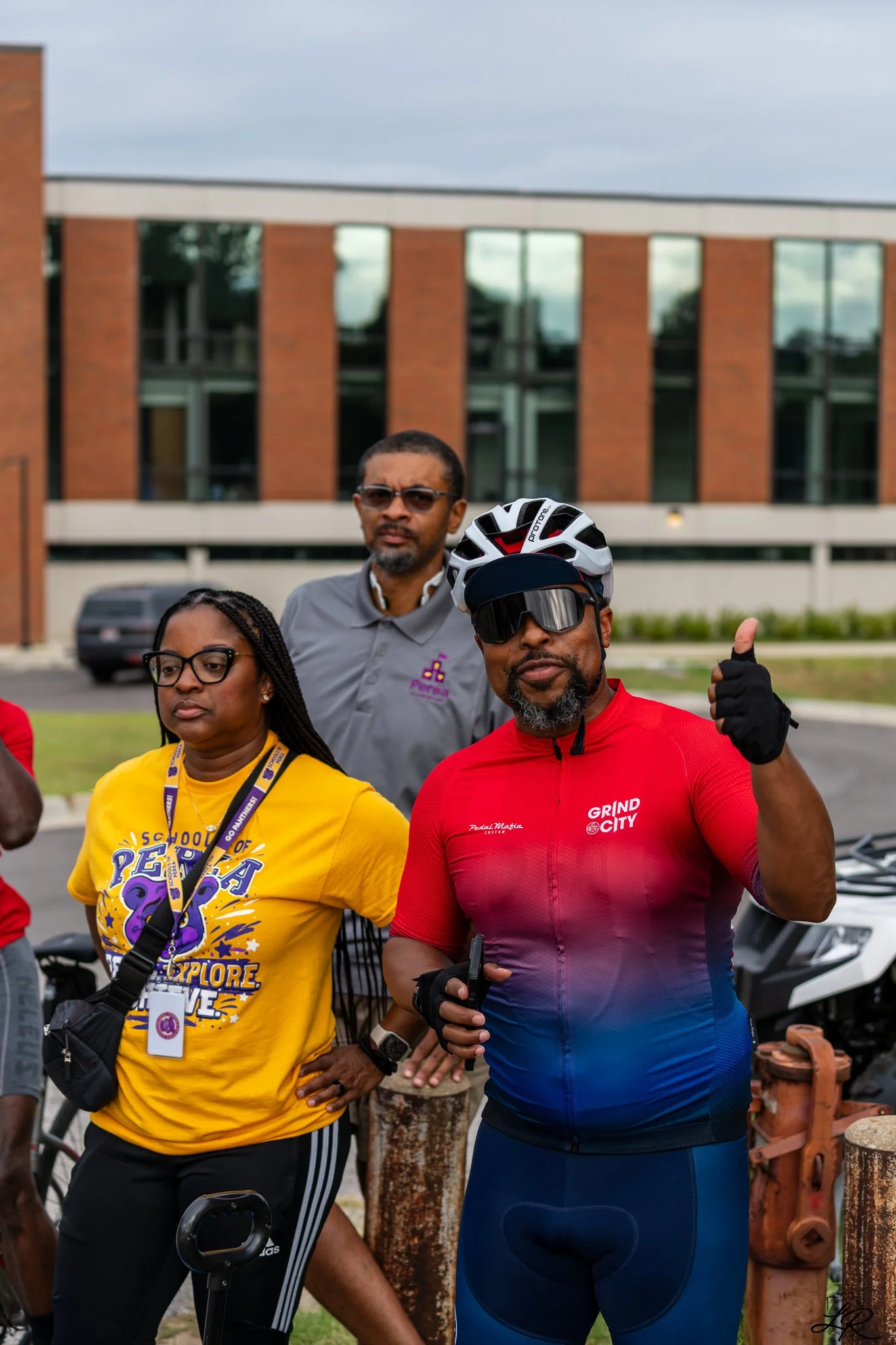 A group of people gathering outdoors, some dressed in cycling gear, with a brick building and cloudy sky in the background.