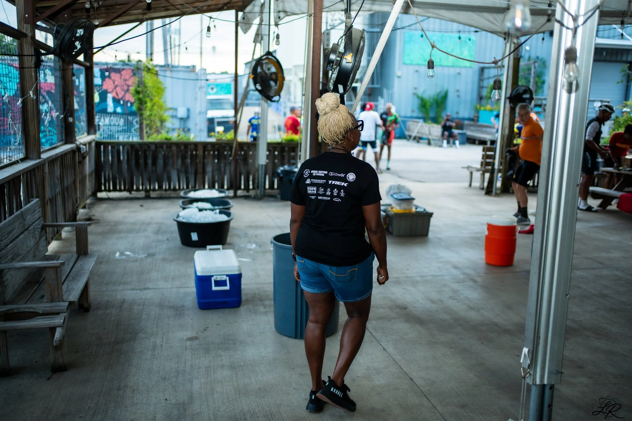 A woman with blonde hair in a bun, wearing glasses, a black t-shirt with logos on the back, and denim shorts, walking on a porch with wooden railing. There are buckets and containers on the floor. In the background, people are outside, and there are 