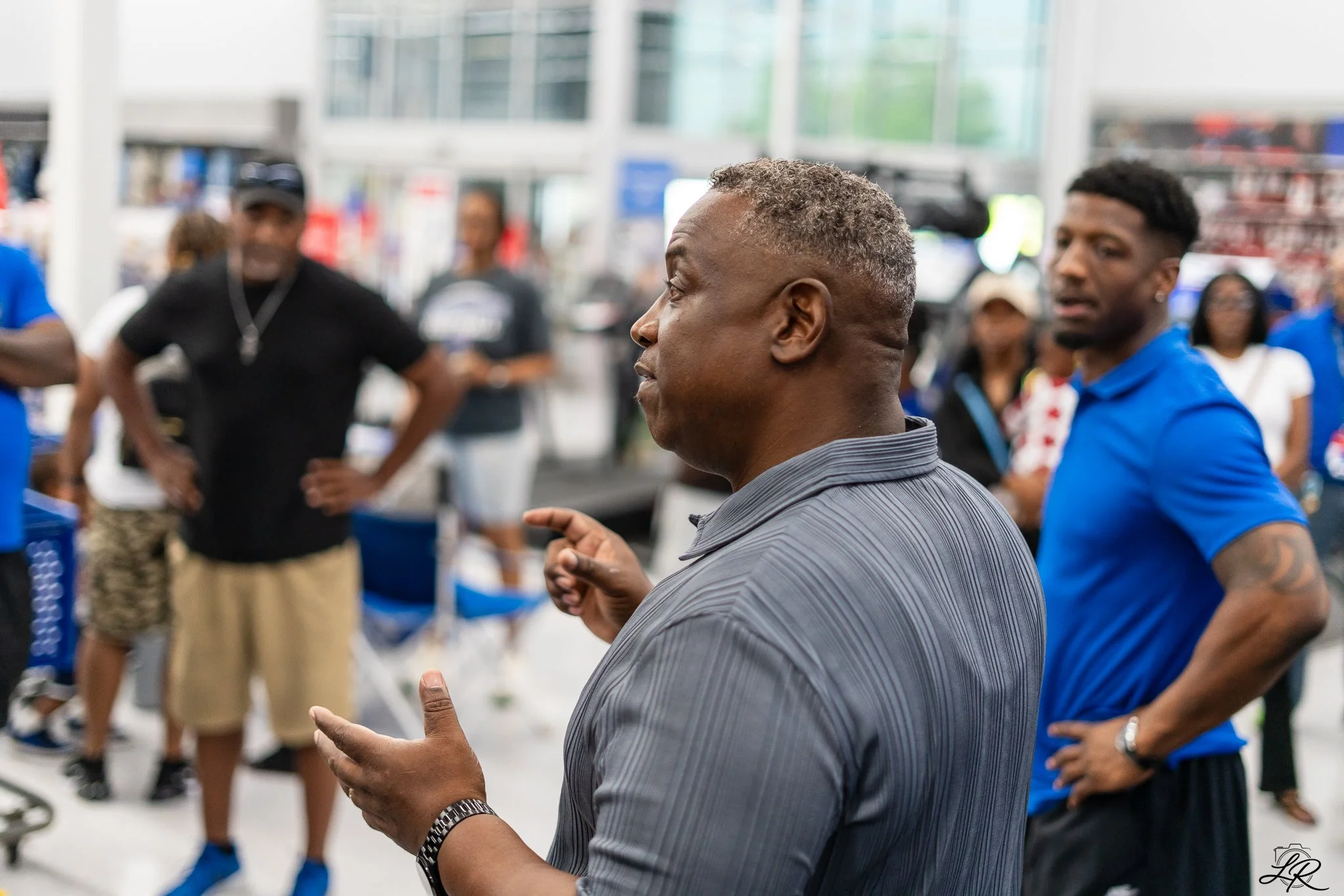 A man in a gray striped shirt and silver watch talking to a group of people at an indoor event.