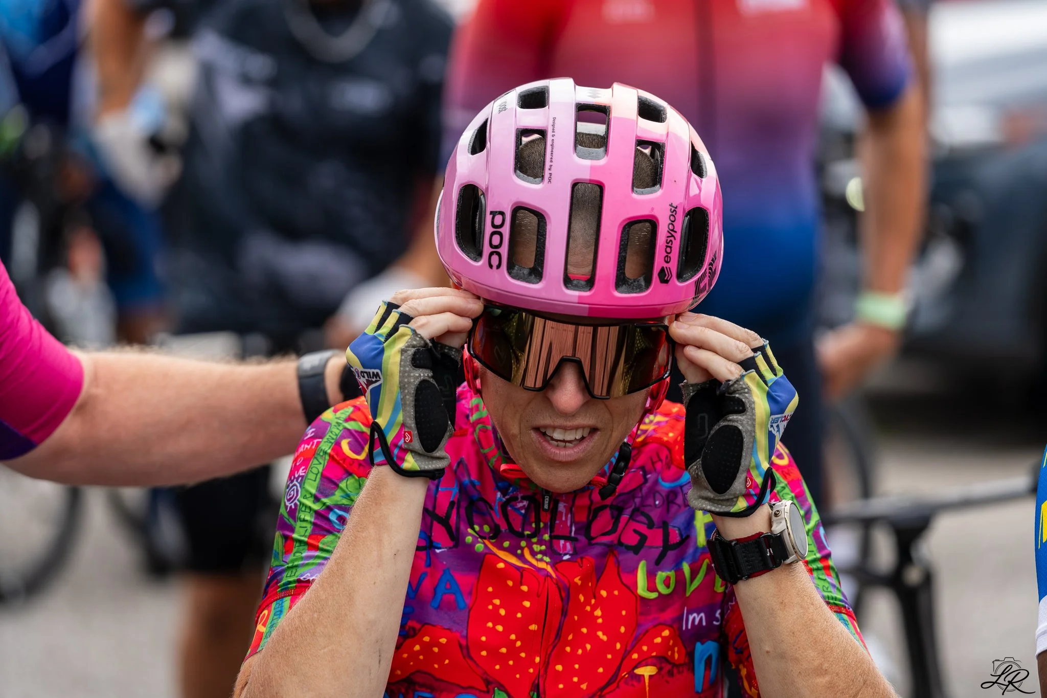 Cyclist adjusting helmet before a race, wearing colorful racing gear and sunglasses, with blurred background of other people.