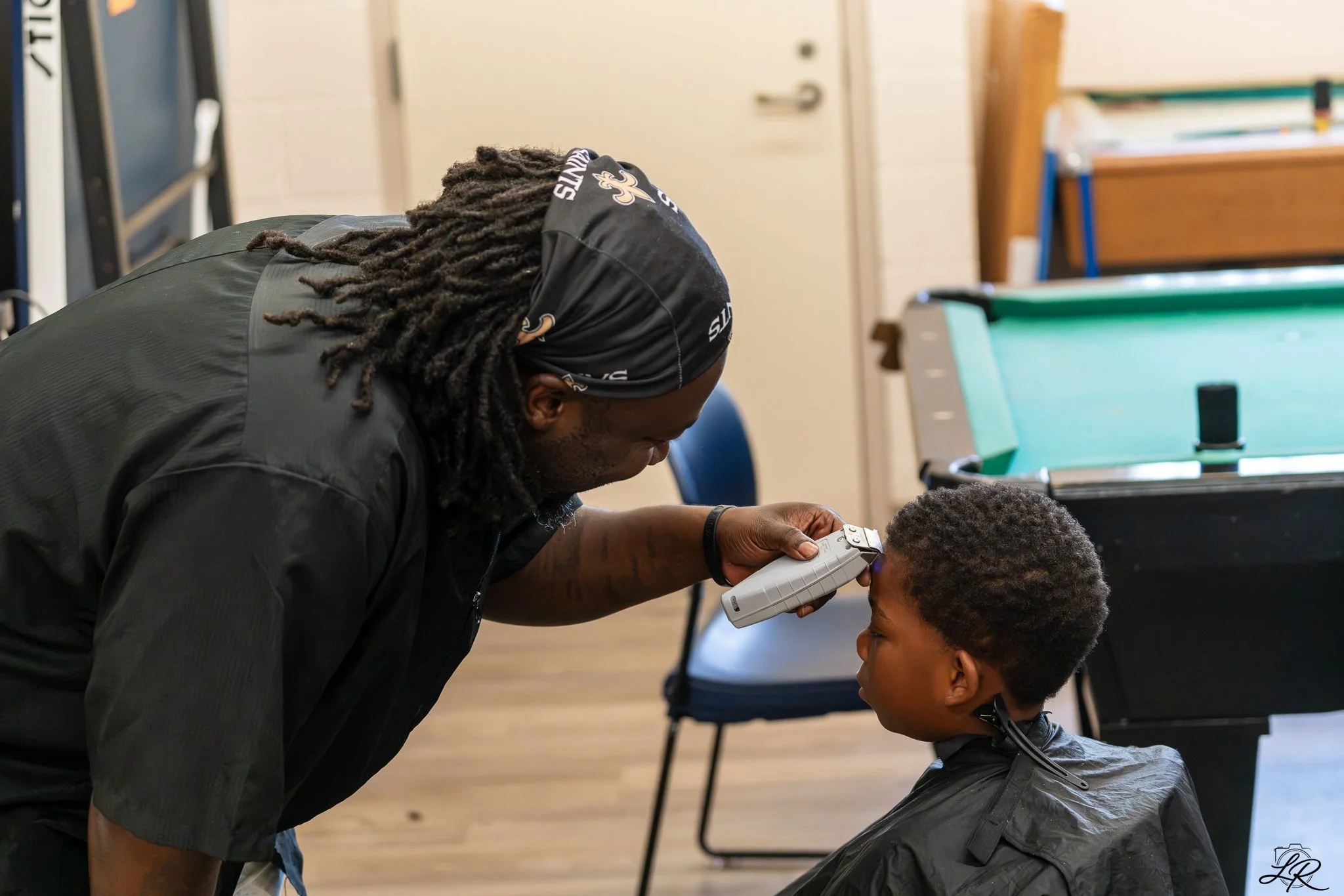 An adult man with dreadlocks wearing a black bandana and black jacket is leaning over a young boy sitting in a barber's chair. The man is holding a handheld device near the boy's temple, possibly a laser tool. In the background, there are chairs, she