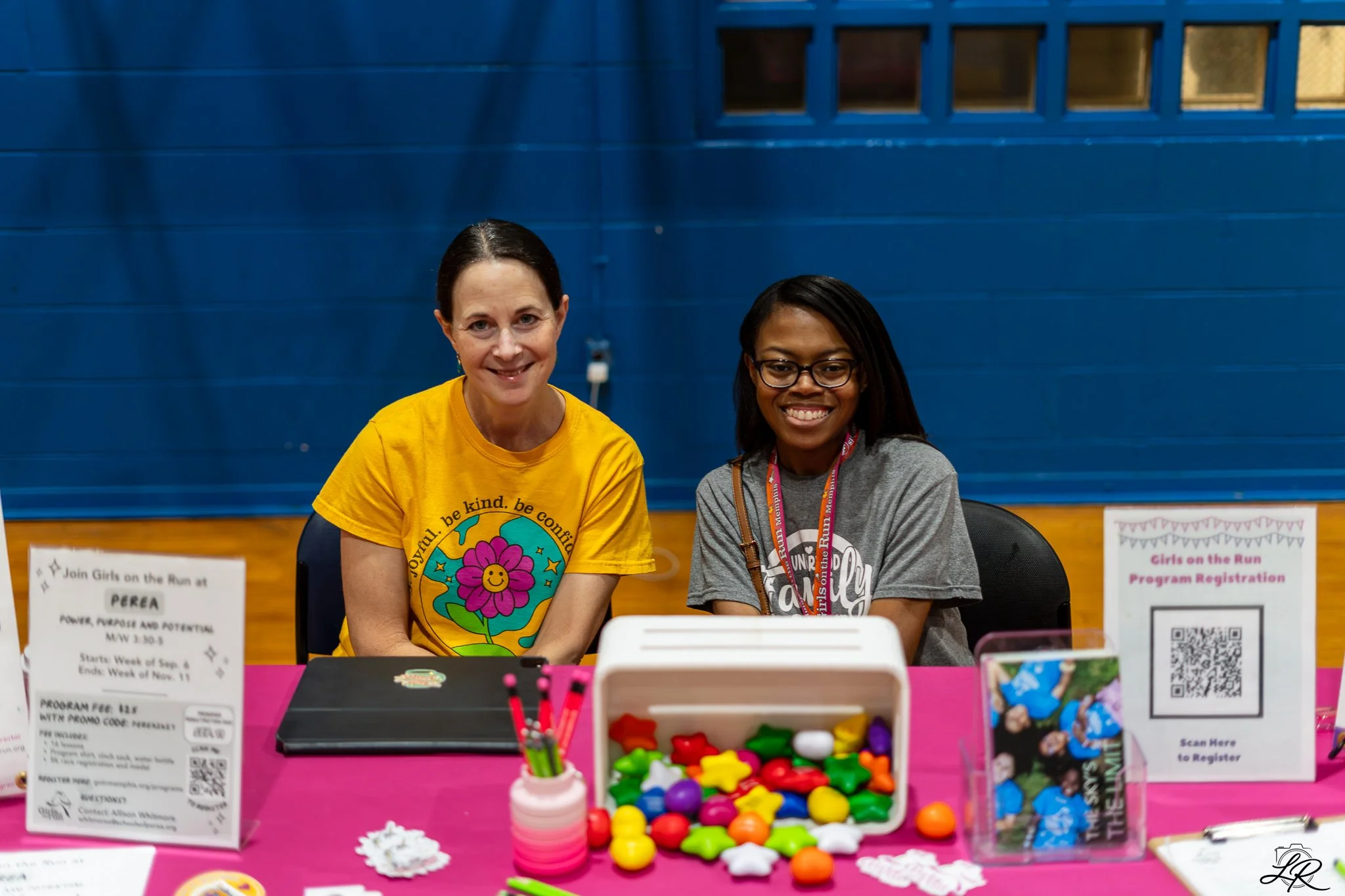 Two women sitting at a pink table with various colorful supplies, smiling, in front of a blue wall with informational signs about a girls' event.
