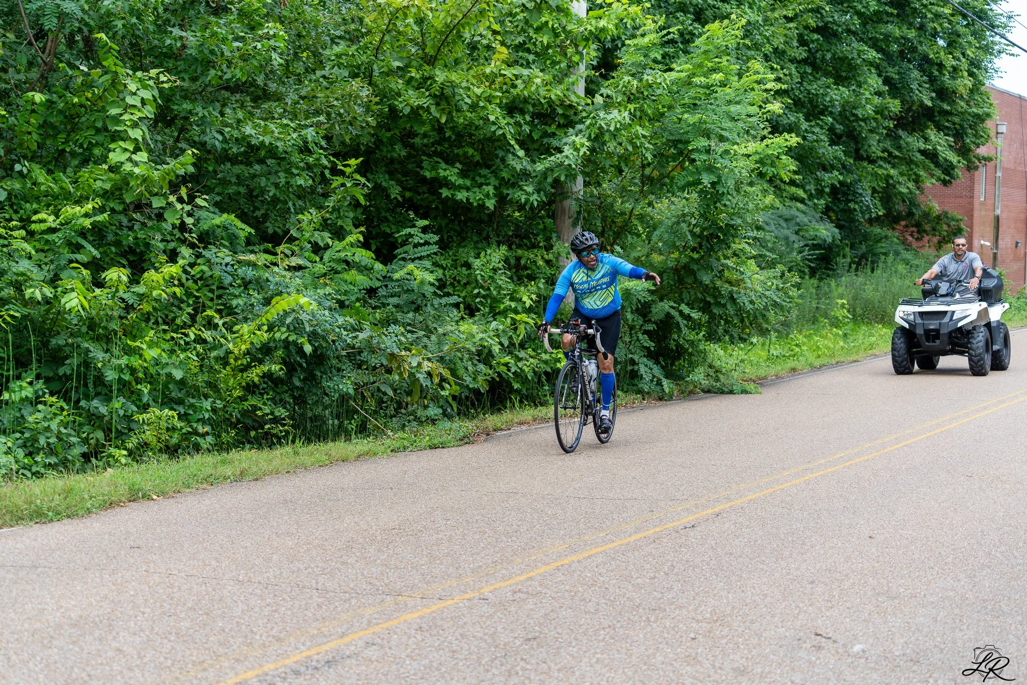 A man riding a bicycle on a paved road next to dense green foliage, with a man driving an ATV behind him.