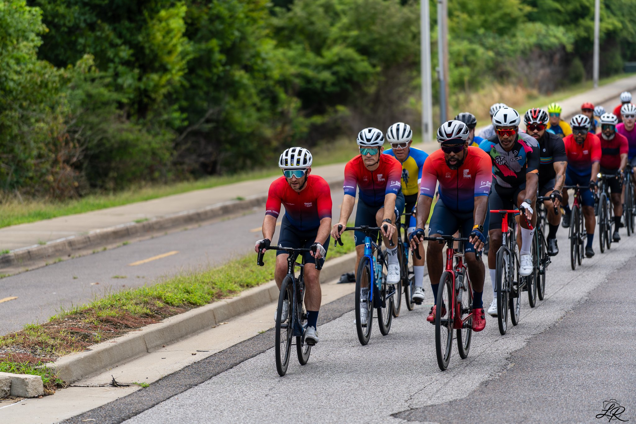 A group of cyclists riding on a paved road surrounded by green trees and bushes.