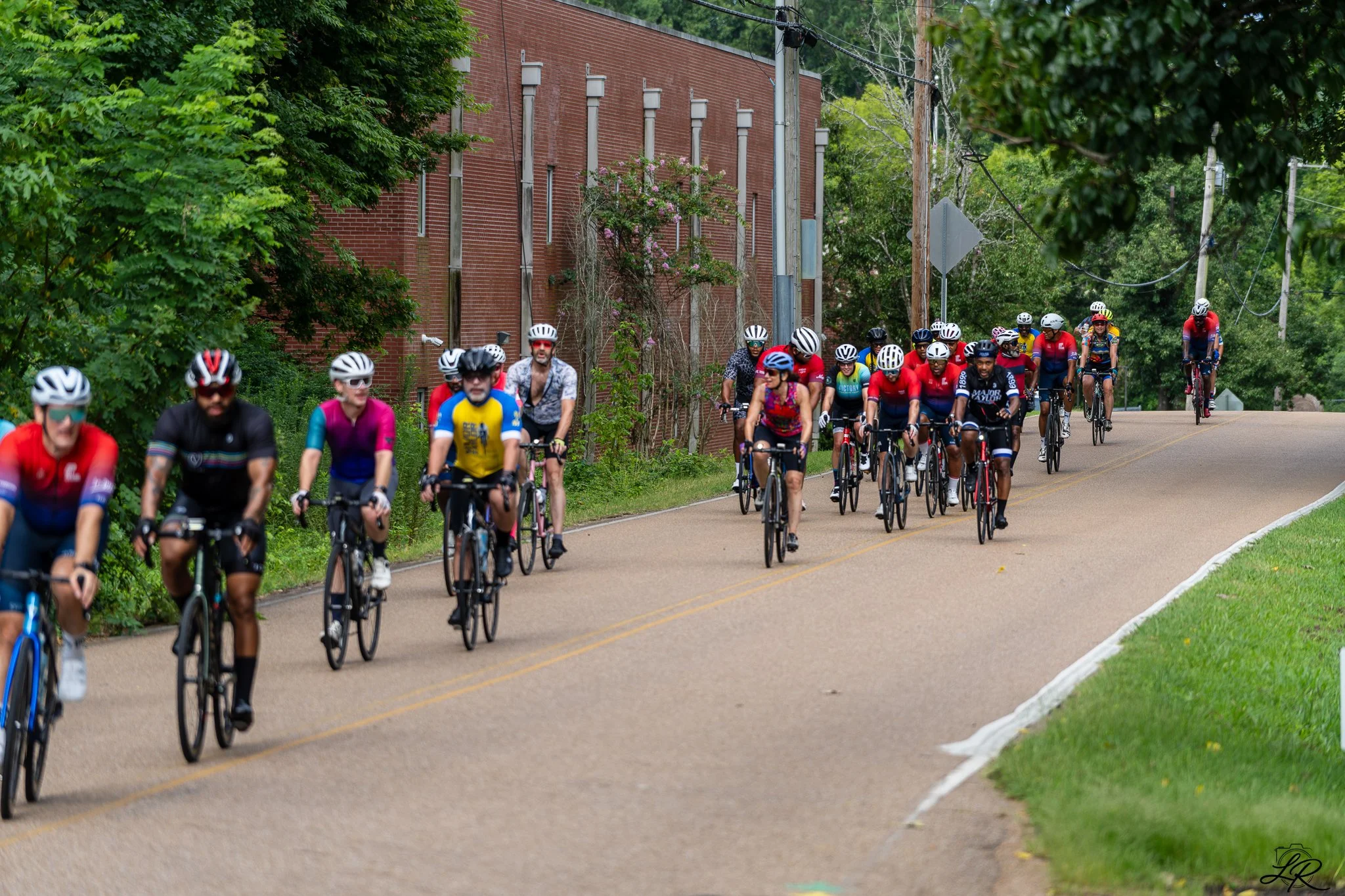 A group of cyclists riding on a paved road surrounded by green trees and a brick building, with utility poles and wires overhead.