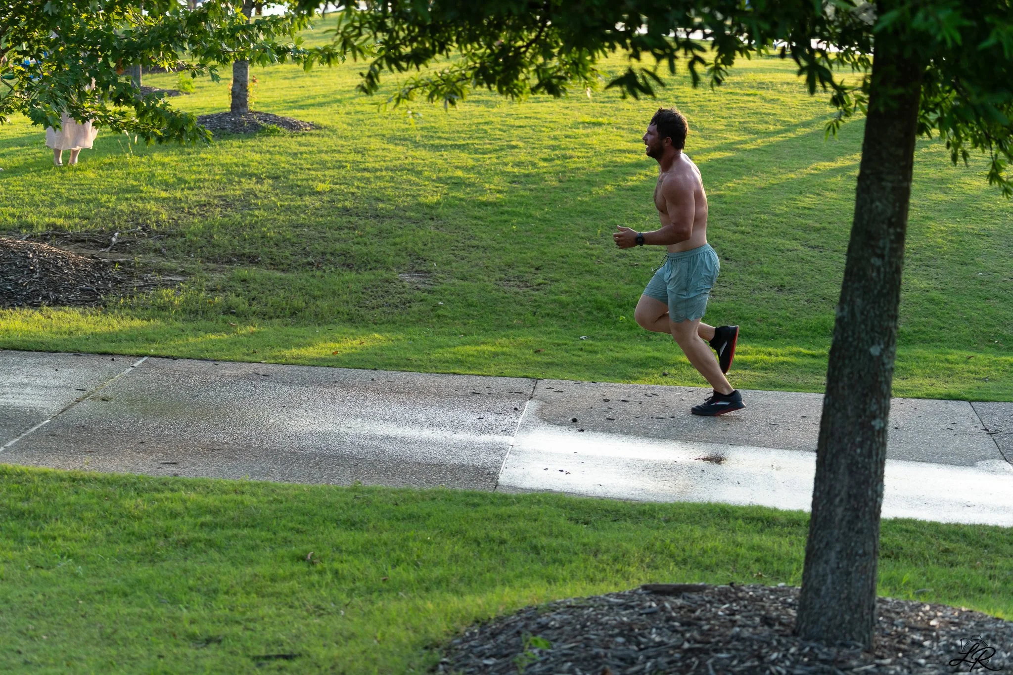 A shirtless man jogging on a wet sidewalk in a park with green grass, trees, and a woman standing under a tree in the background.