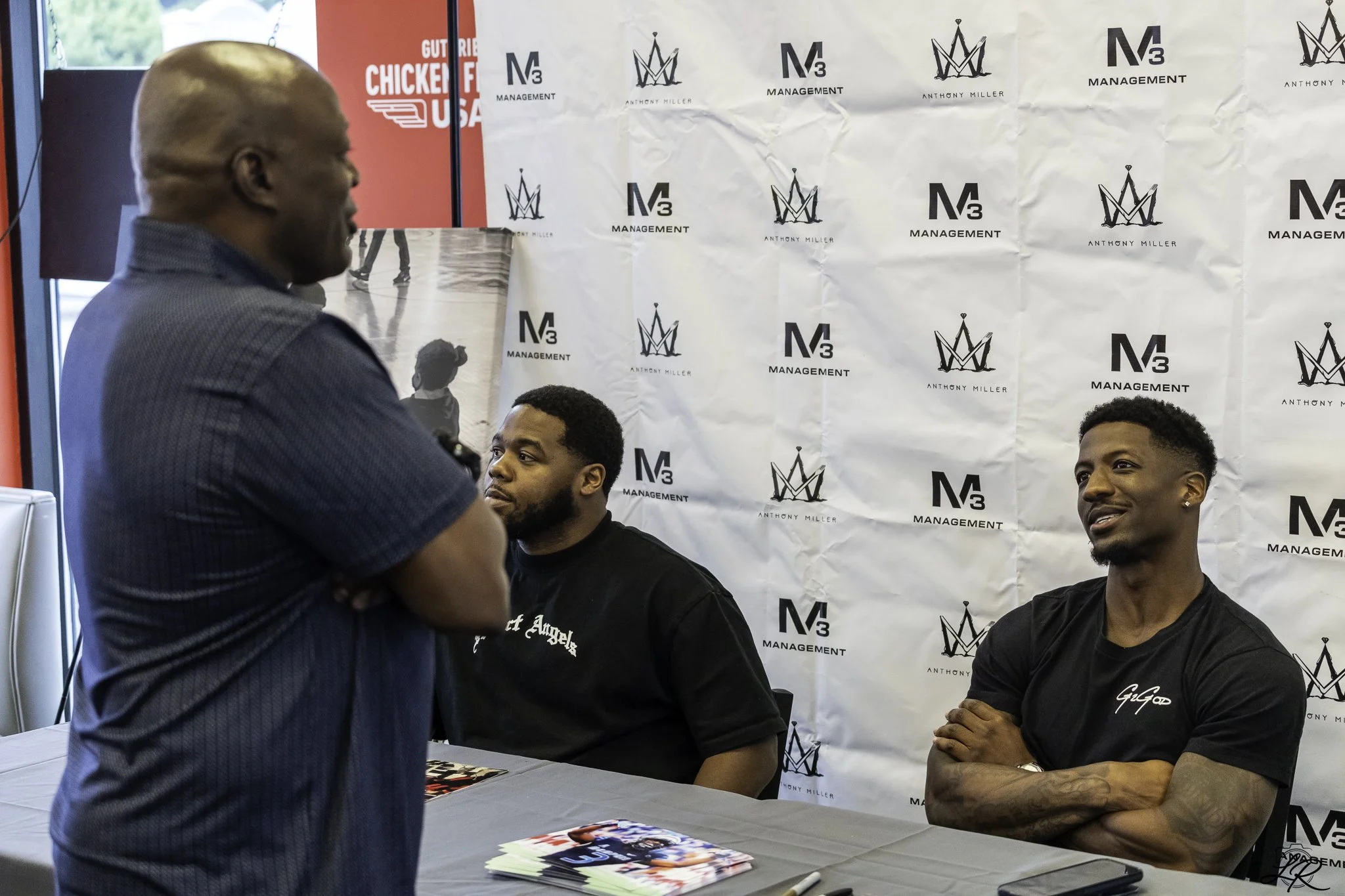 Three young men sitting at a table with a man standing and talking to them, in front of a backdrop with logos that say 'Anthony Miller Management'. The men are wearing black shirts, and there are brochures on the table.