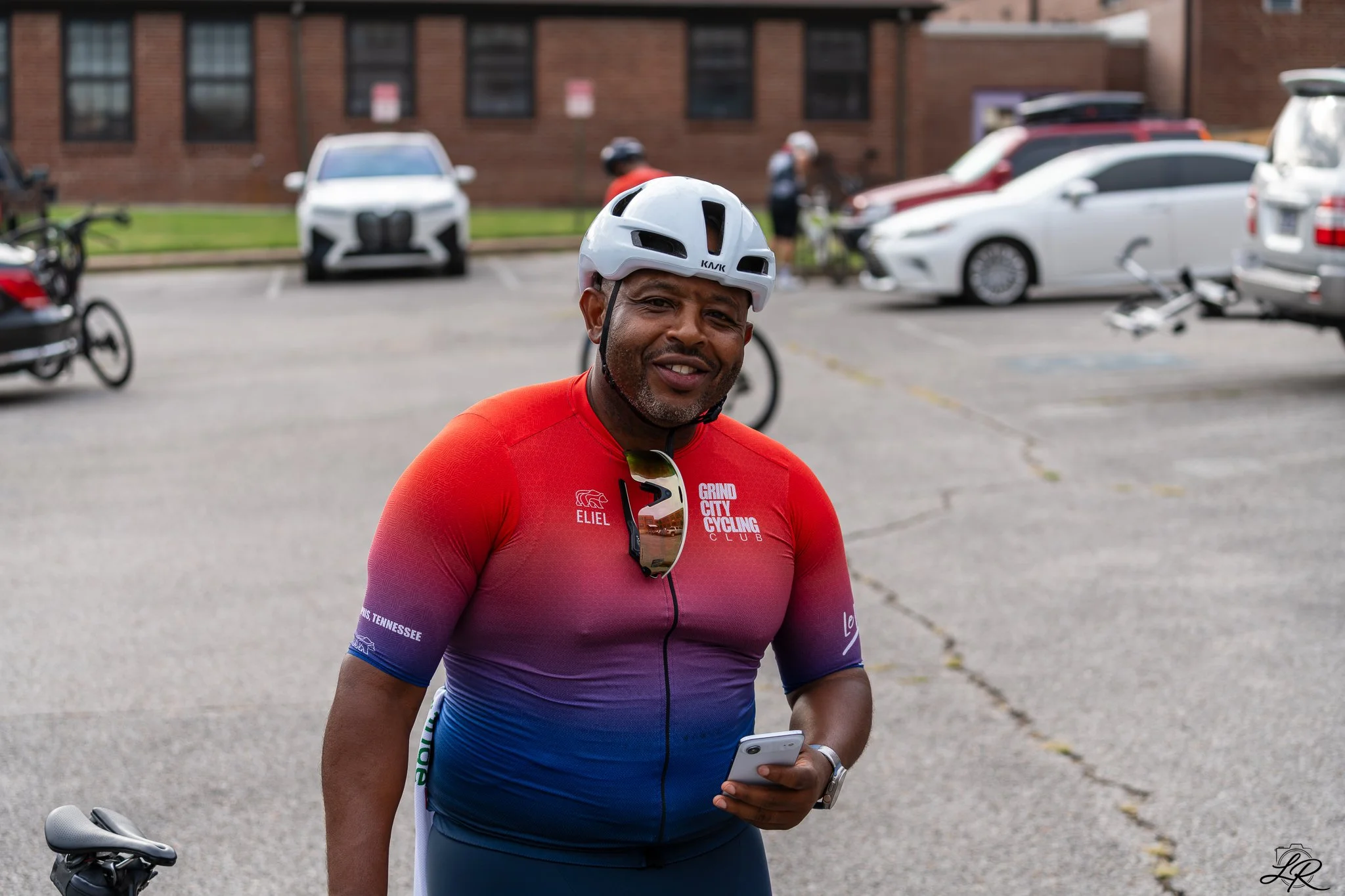 A man wearing a white bicycle helmet and a colorful cycling jersey is holding a smartphone while standing next to a bicycle. Background includes parked cars and a brick building.