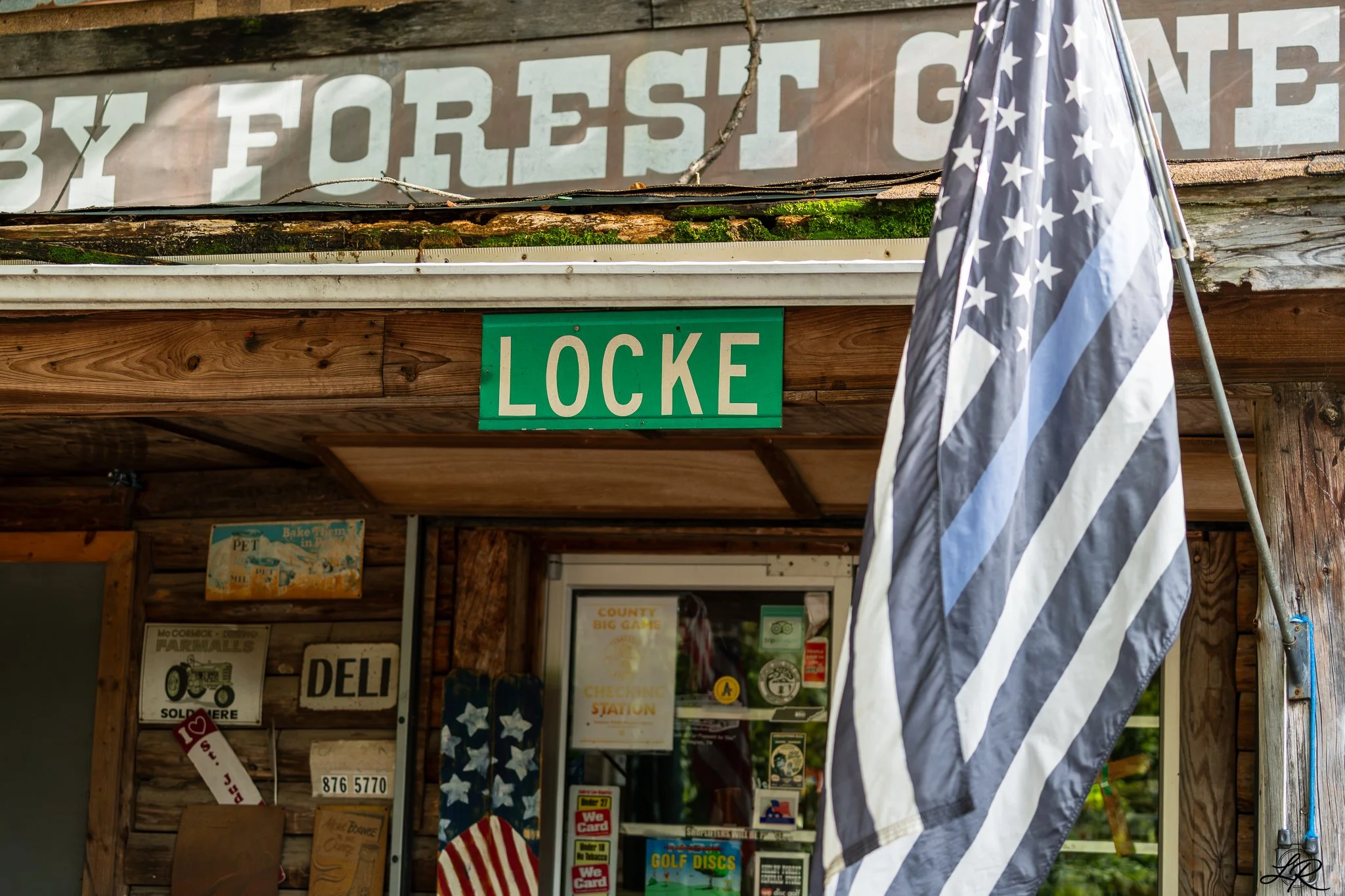 A rustic storefront with a faded sign reading 'BY FOREST' partly covered by a black and white American flag with stars, and a green sign that says 'LOCKE'. Various vintage posters and signs are visible on the wooden wall inside.
