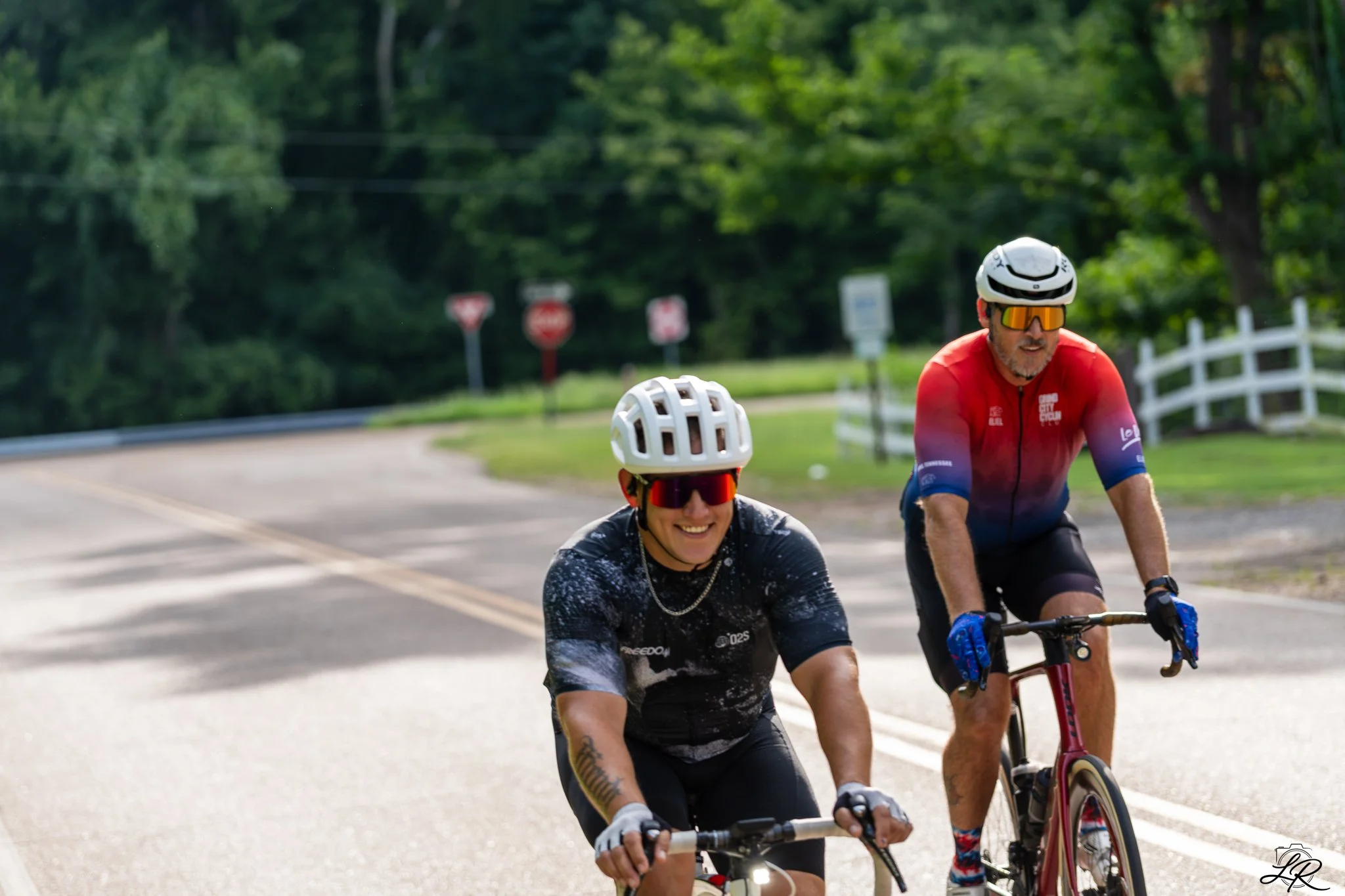 Two cyclists, a woman and a man, smiling and riding bikes on a road with trees and stop signs in the background.