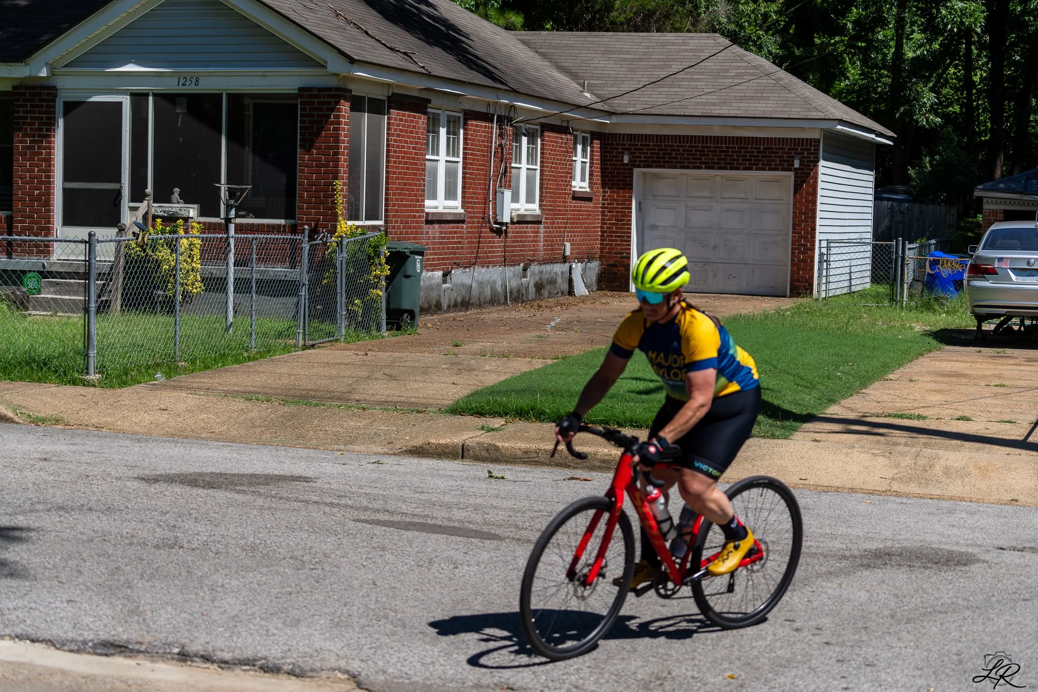 A person wearing a yellow and blue cycling jersey, yellow helmet, and sunglasses riding a red bicycle on a residential street. In the background, there is a brick house with a white garage door, a chain-link fence, and green trees.
