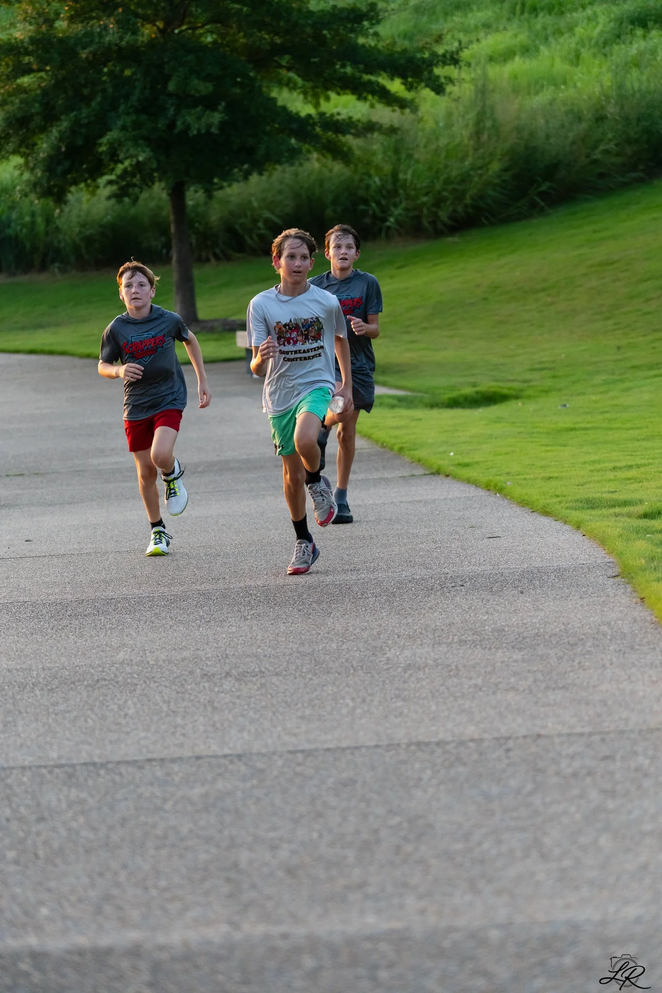 Three boys running on a paved path in a park with green grass and trees.
