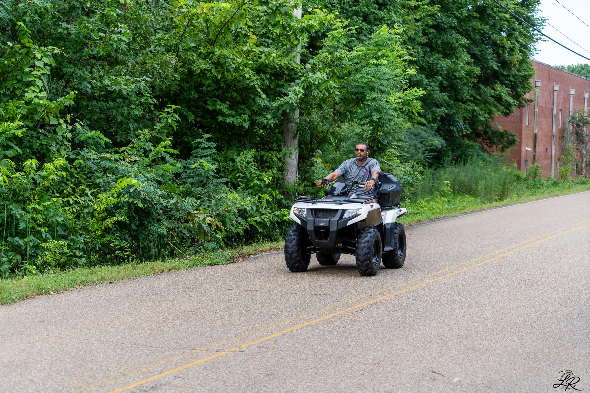 A man driving a quad bike with a top box along a road next to green trees and a brick building.
