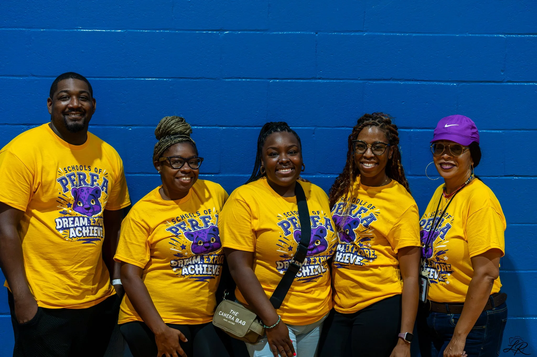 Group of six diverse people standing in front of a blue wall, wearing matching yellow T-shirts with purple and white text and logo for a school event. They are smiling and posing for the photo.