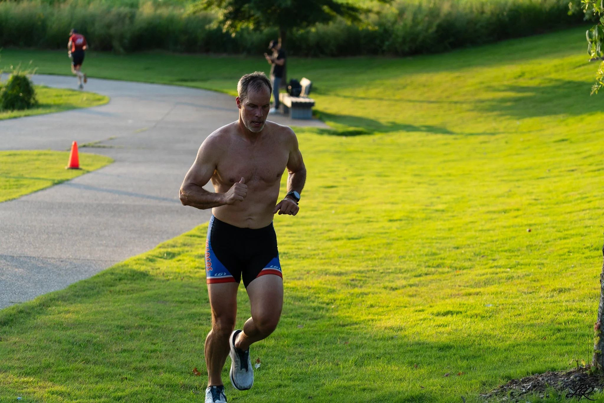 A shirtless man running outdoors on a grassy pathway in the late afternoon or early evening, with two people in the background and trees.
