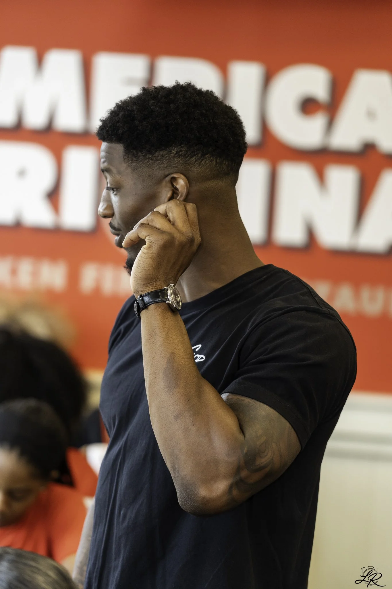 Side profile of a young man with curly hair and a beard, wearing a black T-shirt and a wristwatch, touching his ear, with a red background that has large white text.