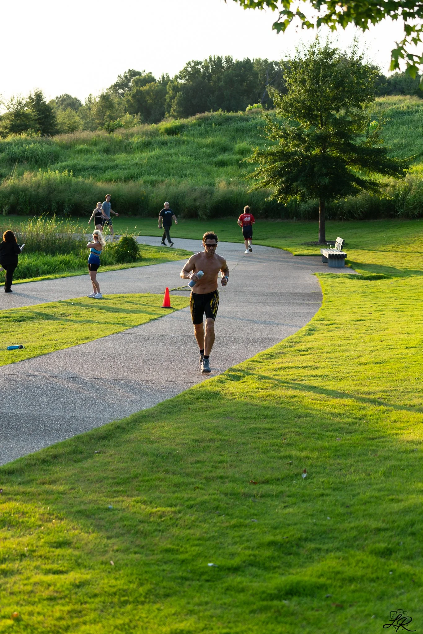 A park with a paved pathway, green grass, trees, and several people jogging, walking, and standing, enjoying the outdoor scene in the late afternoon or early evening.