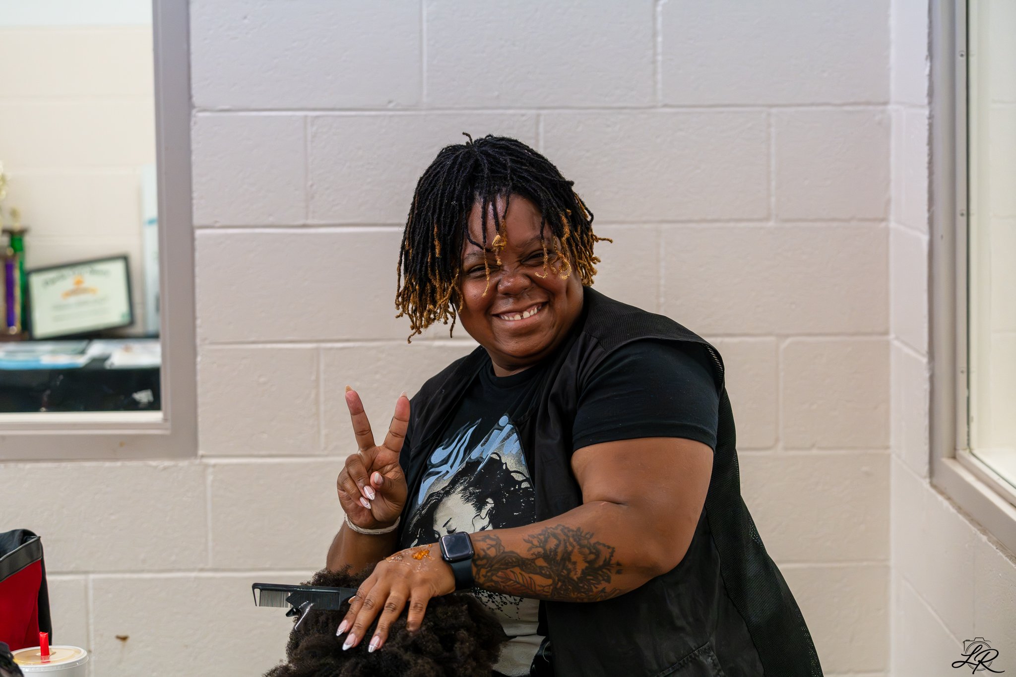 A woman with short dreadlocks is smiling and making a peace sign while sitting at a table, with a person in front of her. The setting appears to be indoors with a gray brick wall in the background.