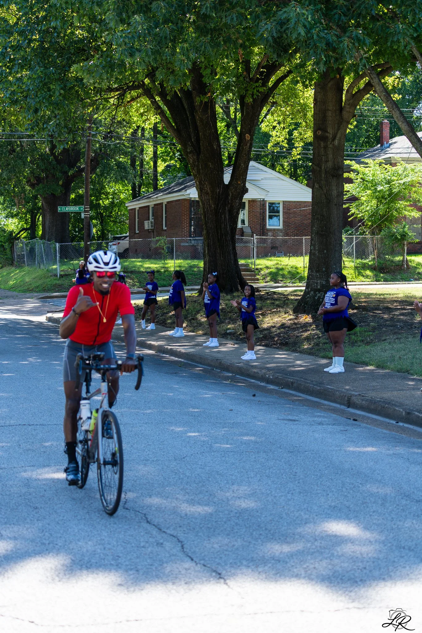 A cyclist in a red shirt and helmet riding a bicycle on a street with a group of young girls in blue uniforms standing on the sidewalk under trees. Houses and a street sign reading "N Claybrook St" are visible in the background.
