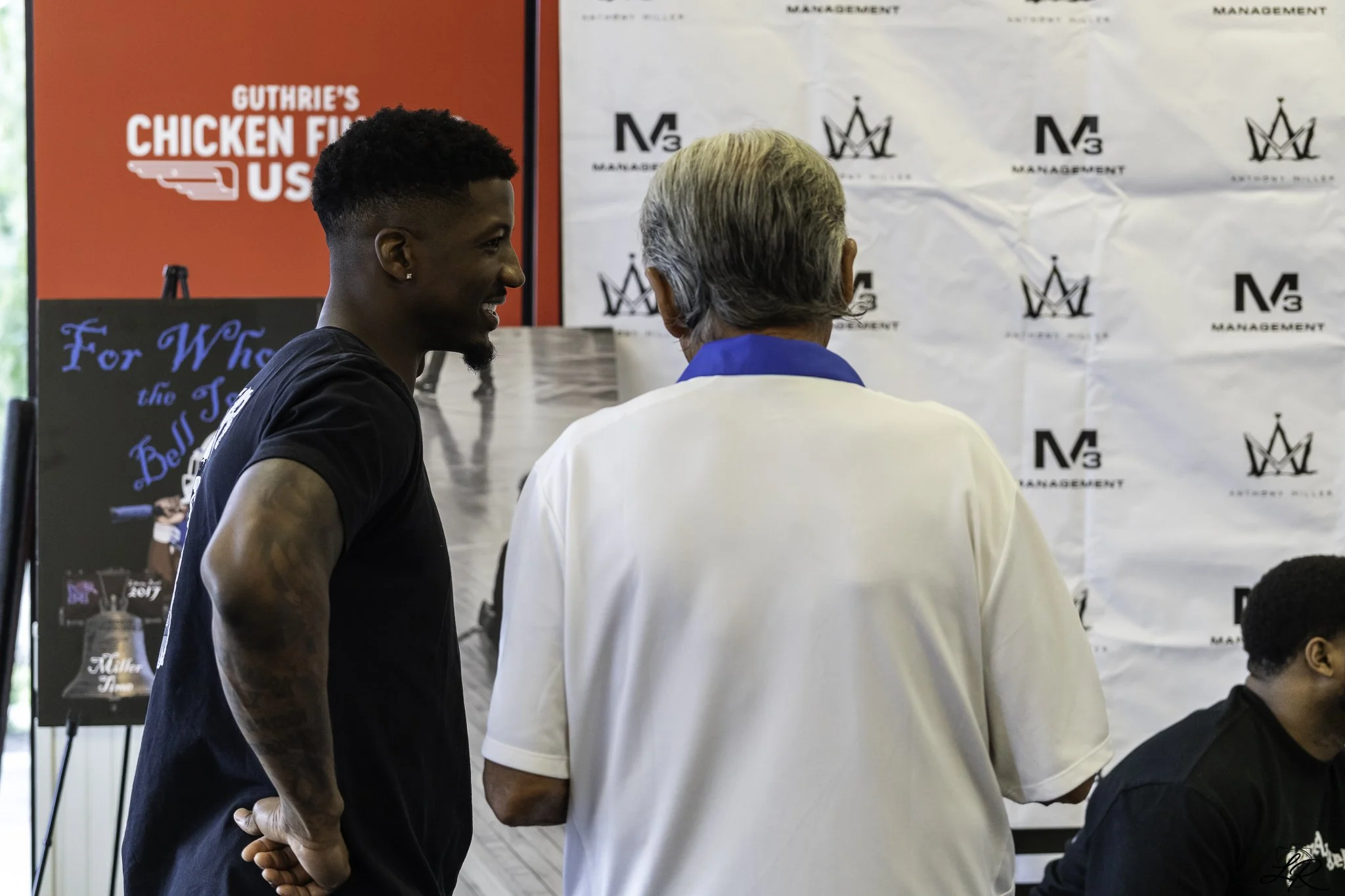 Two men are engaged in conversation at a public event, with one facing away from the camera and the other smiling and looking to their right. The background features banners and signs, including one with red and white text reading "Guthrie's Chicken 