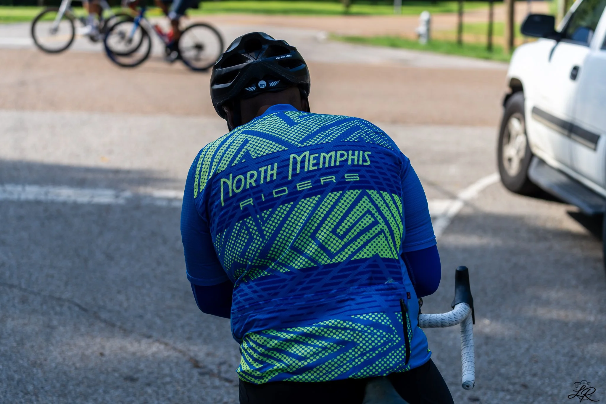A person dressed in a bright cycling jersey with the words "North Memphis Riders" on the back, wearing a black helmet, and holding a bicycle, with two children riding bicycles blurred in the background.