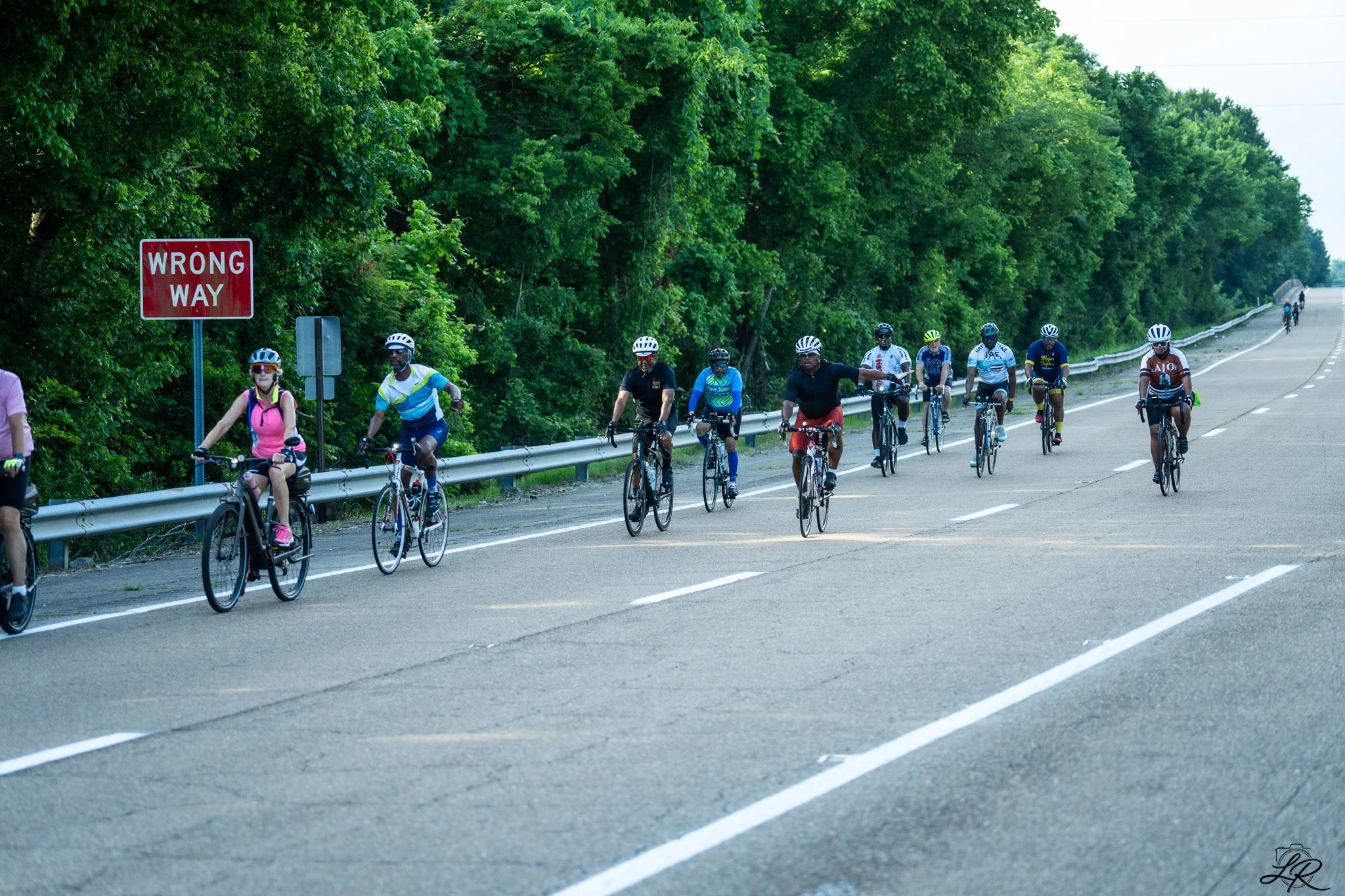 A group of cyclists riding on a road surrounded by green trees, with a red "Wrong Way" sign on the left side.