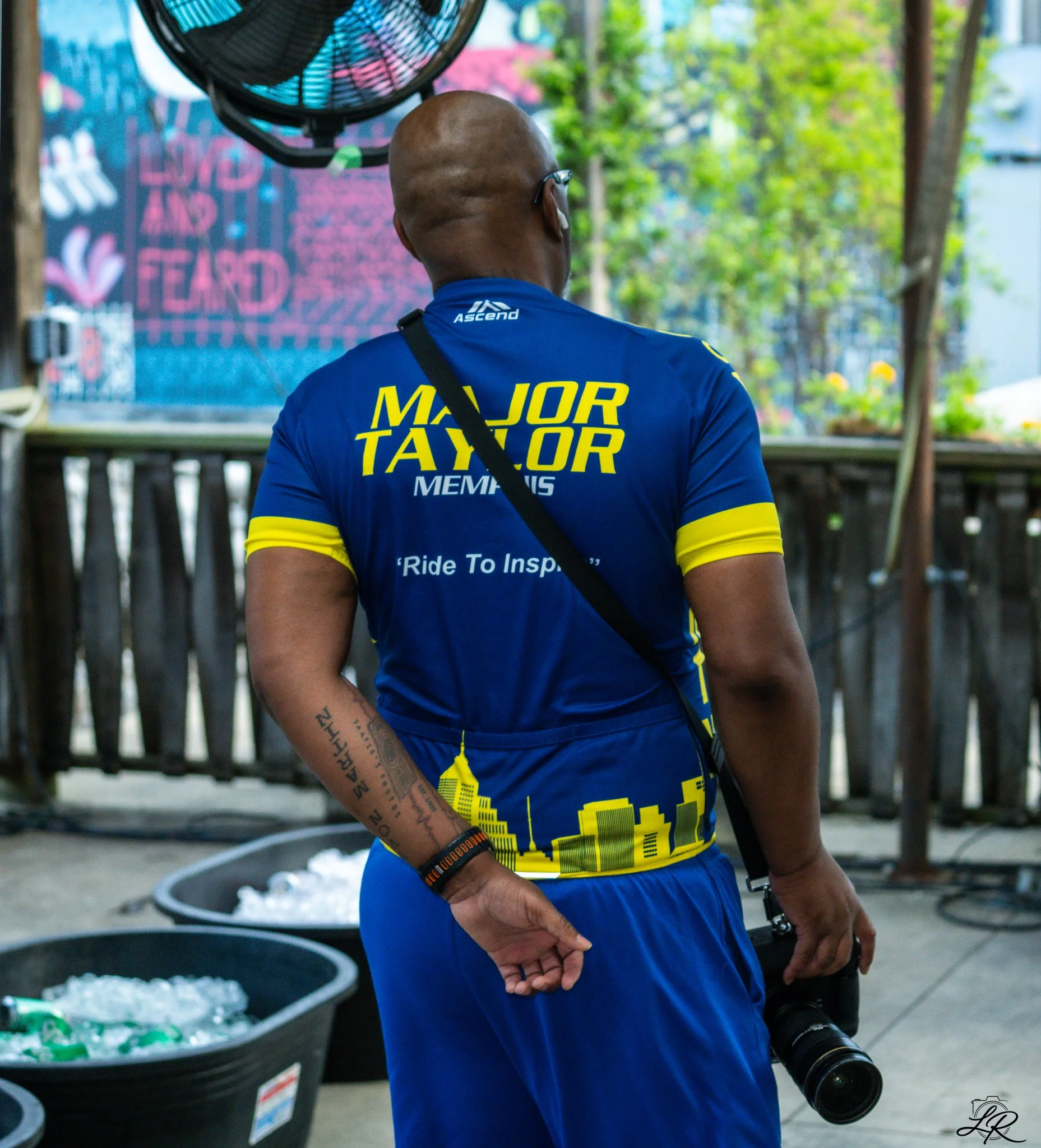 Back view of a man wearing a blue T-shirt with yellow text that reads "Major Taylor Memphis" and a black strap across his shoulder, standing outdoors near containers of ice and a large cross-shaped plant arrangement, holding a camera with a long zoom