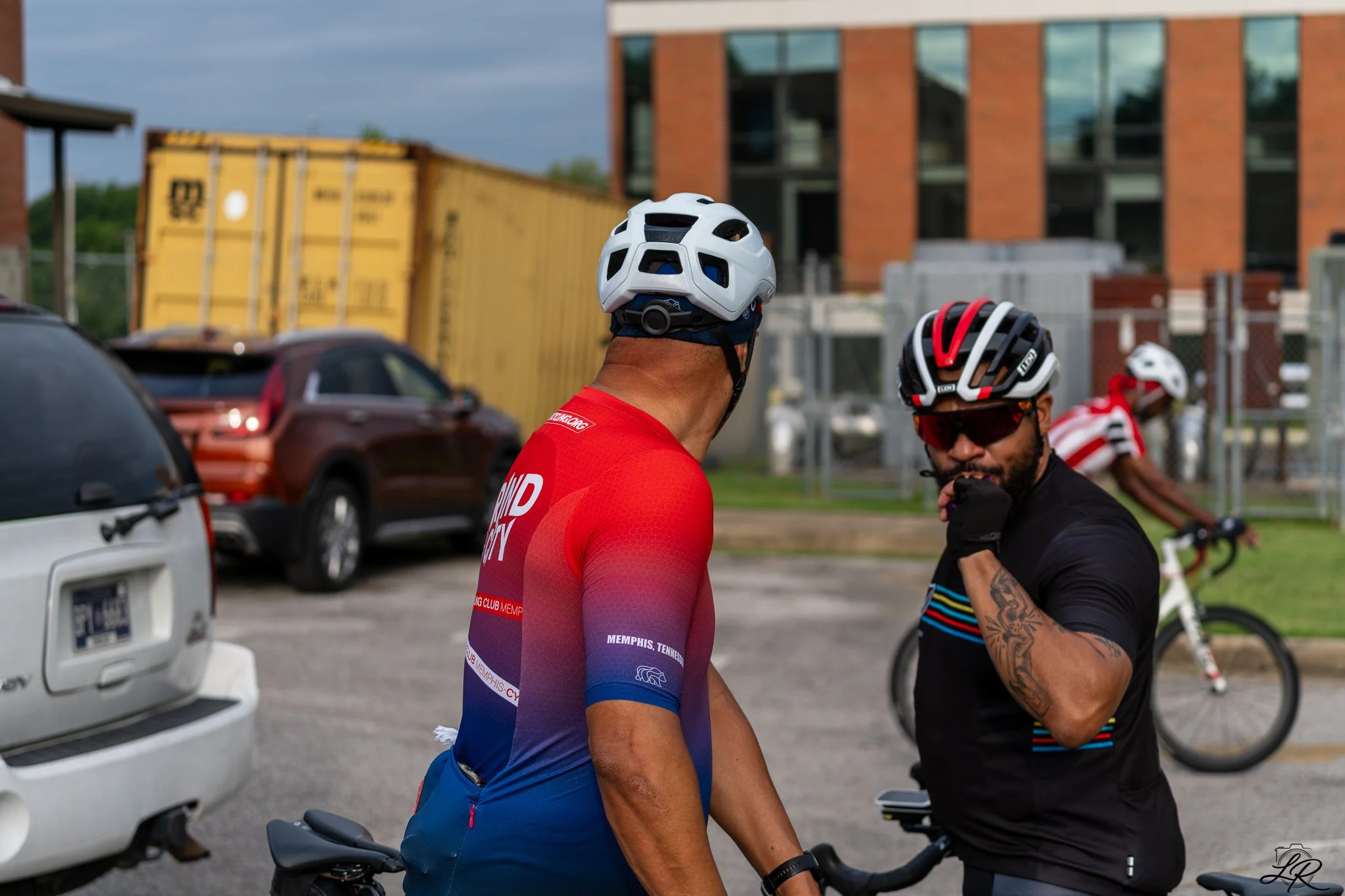 Two cyclists wearing helmets and colorful cycling gear engaged in conversation in a parking lot, with cars and a modern brick building in the background.