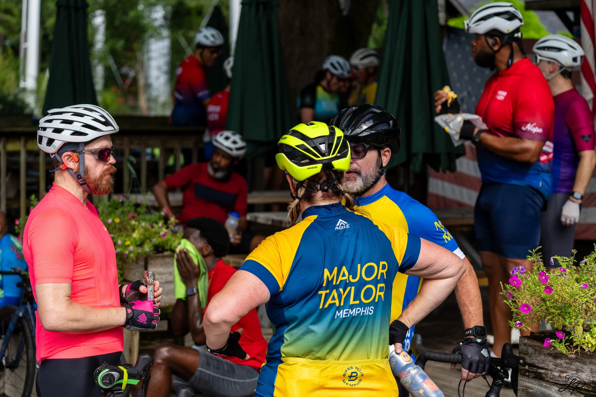 Group of cyclists wearing helmets and colorful cycling apparel gathered in a park, with some holding snacks or drinks, amidst trees and plants.