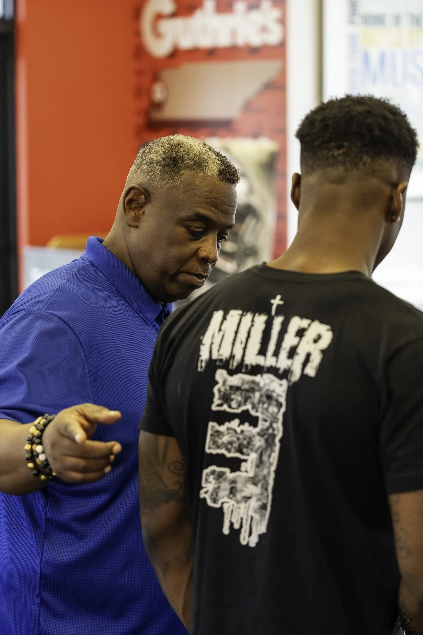 A man in a blue shirt with short hair, some blonde highlights, and bracelets on his wrist, appears to be talking to a younger man with a fade hairstyle and a black t-shirt with white graphics on the back, inside a store or restaurant with red walls a