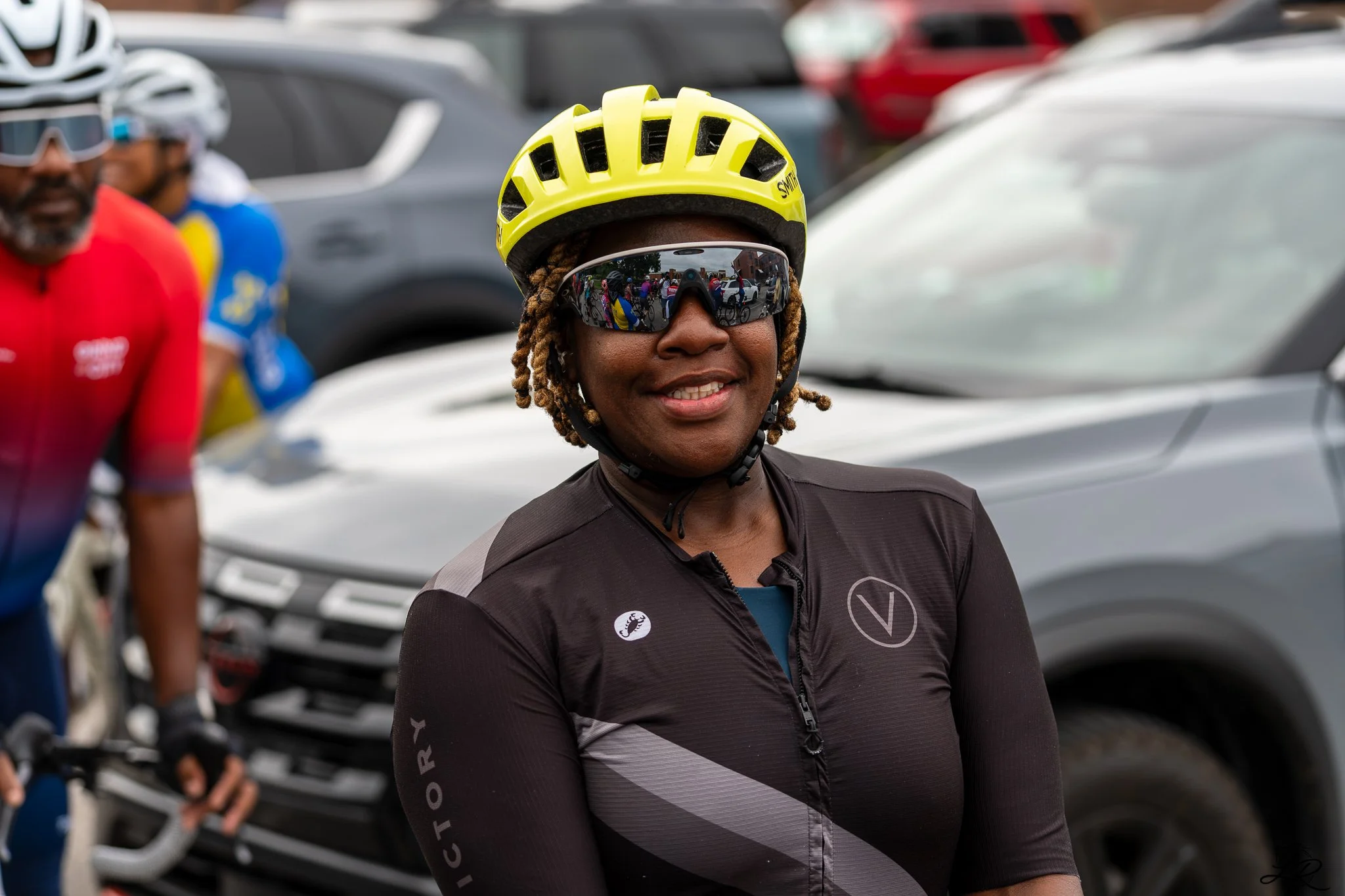 Smiling woman wearing a yellow bike helmet and reflective sunglasses, standing in front of parked cars during a cycling event.